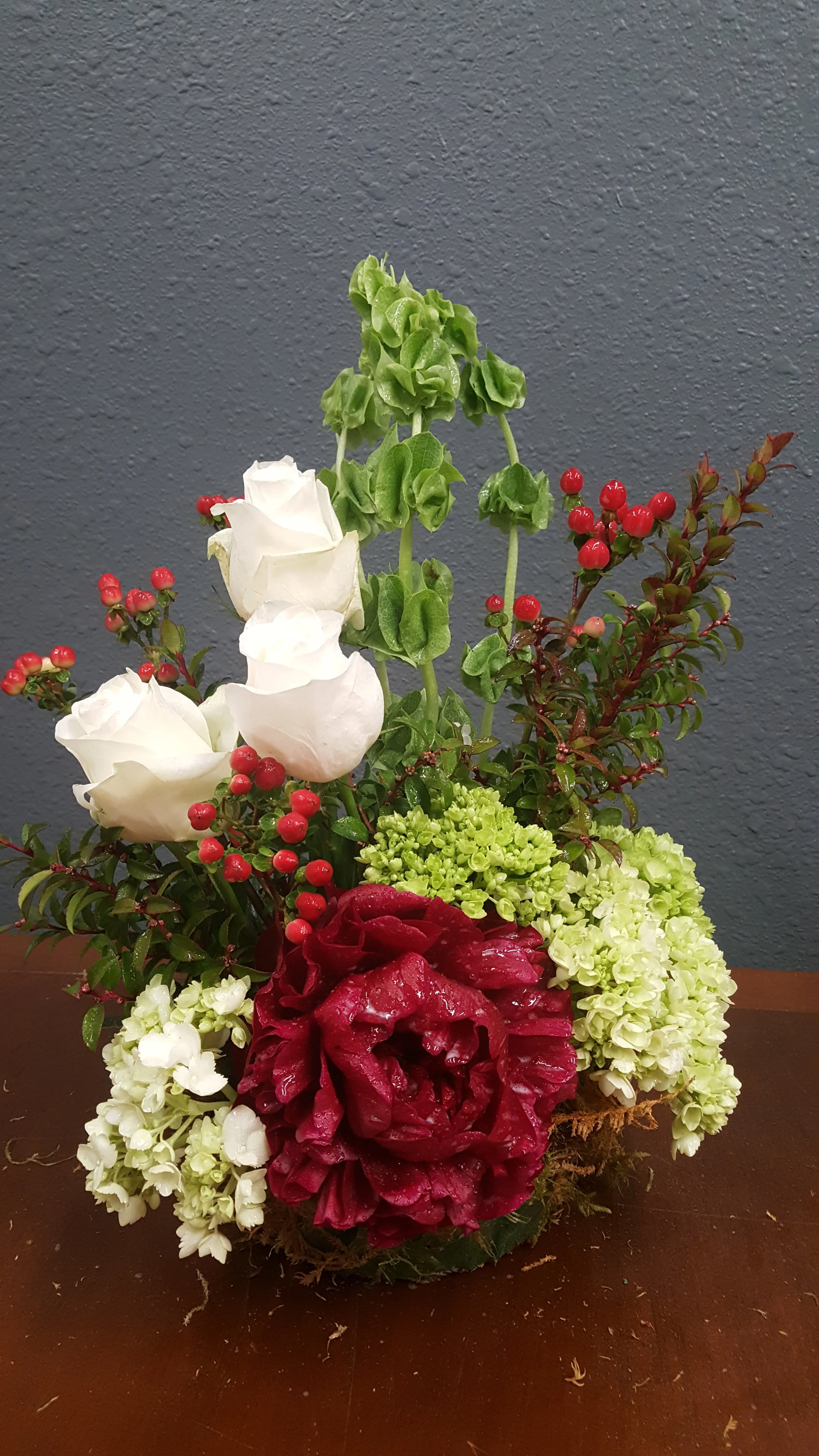 The eye is drawn to the deep crimson peony, a stunning flower in this artful memorial tribute. Roses, hydrangeas, bells of Ireland and hypericum berries complete this beautiful floral display.