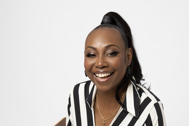  Headshot of a confident black woman laughing, with her hair in a very high ponytail, wearing a black and white striped top with a gold jewellery  