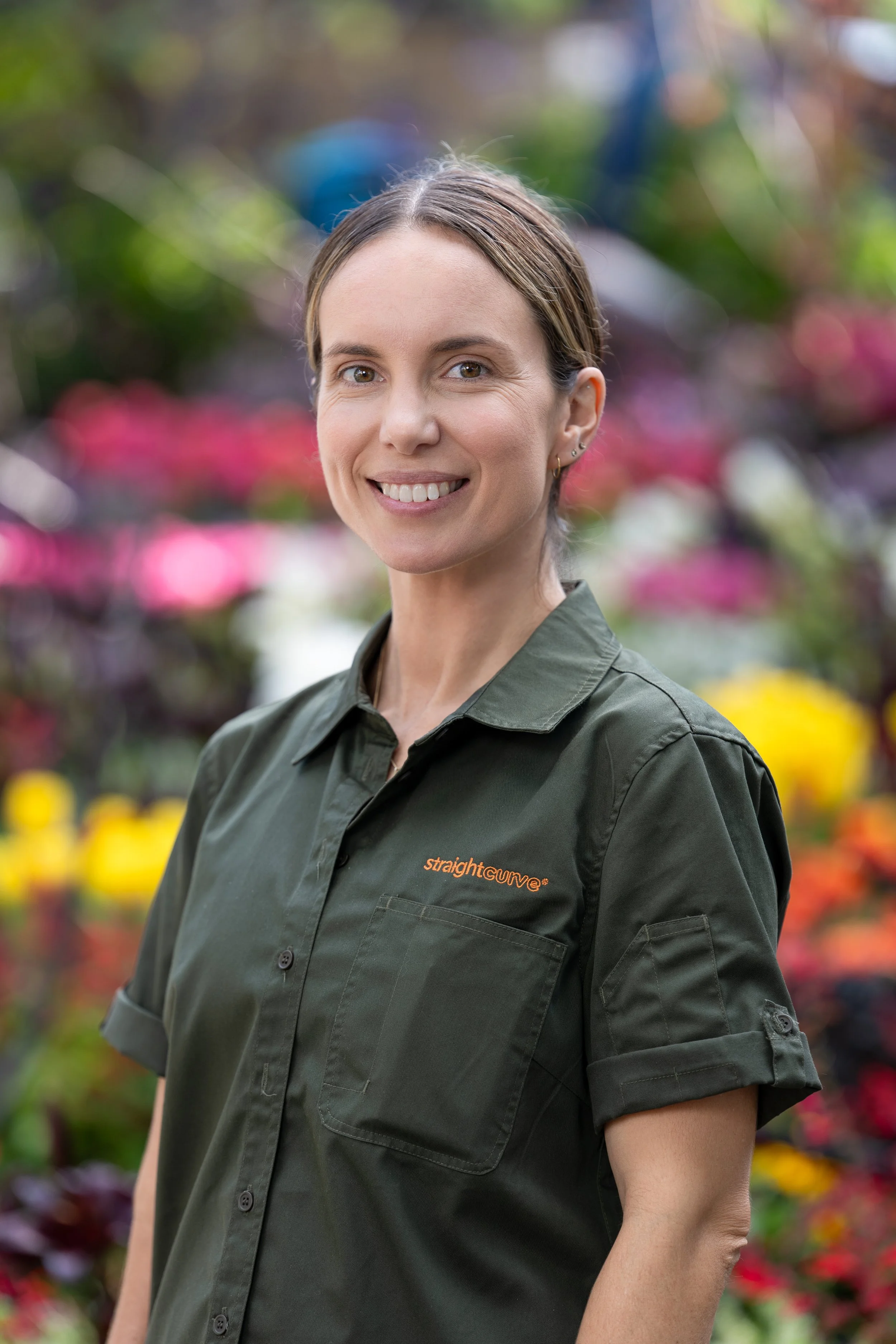 A woman with light skin and brown hair smiling outdoors in front of colorful blurred flowers, wearing a dark green shirt with the logo 'straightcurve'.