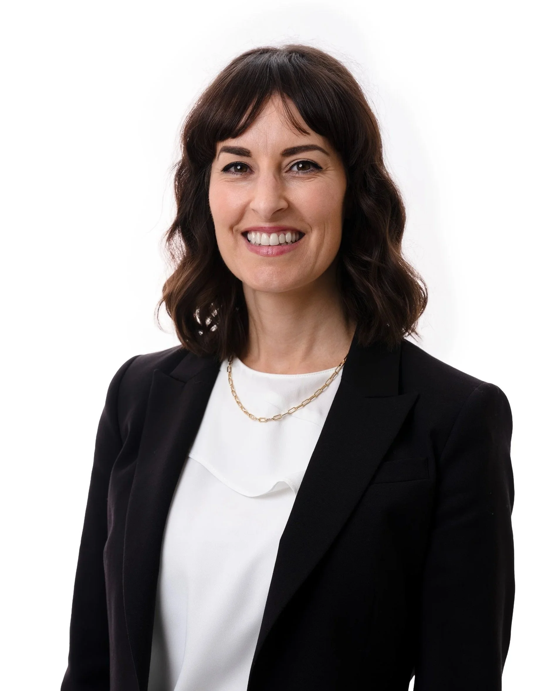A smiling woman with shoulder-length dark brown hair, wearing a white top and a black blazer, accessorized with a gold chain necklace, against a plain white background.