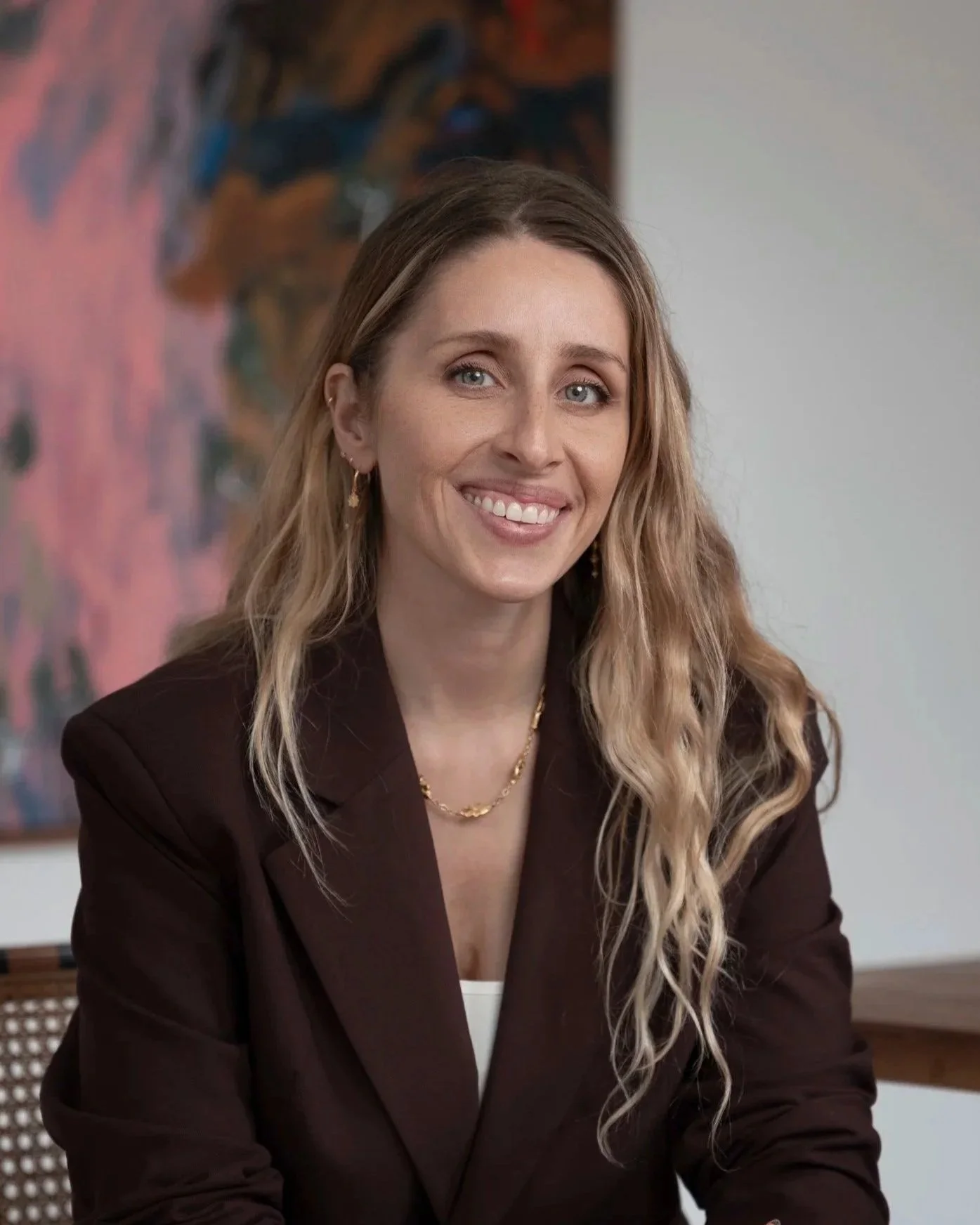 A woman with long wavy blonde hair, wearing a dark blazer and jewelry, is smiling while sitting indoors with a colorful abstract painting in the background.