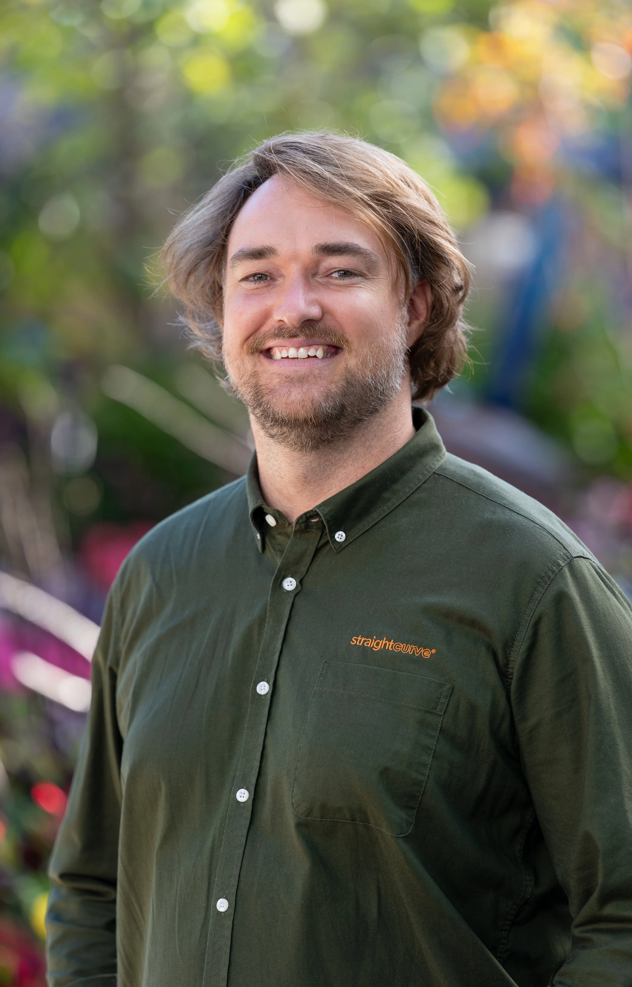 A man with shoulder-length light brown hair, a light beard, and blue eyes smiling outdoors. He's wearing a green button-up shirt with the words 'straightcurve' embroidered on the pocket. The background features blurred trees and colorful foliage.