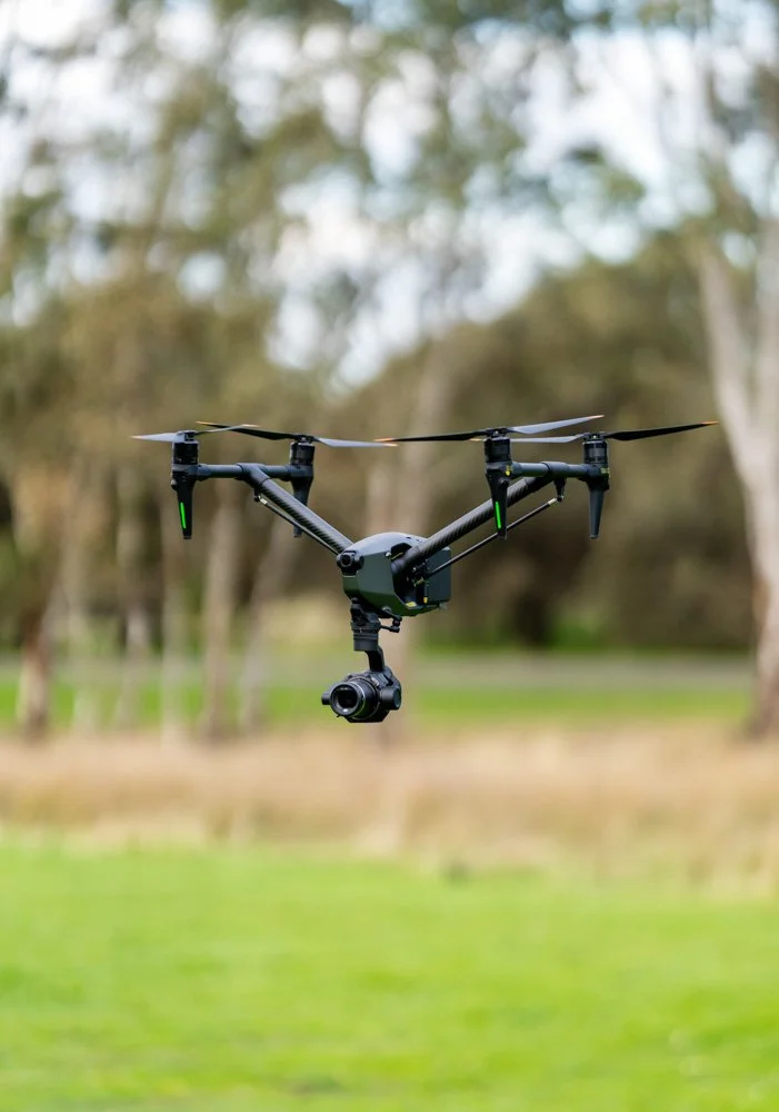 A drone with a camera attached is flying outdoors in a park or forested area.
