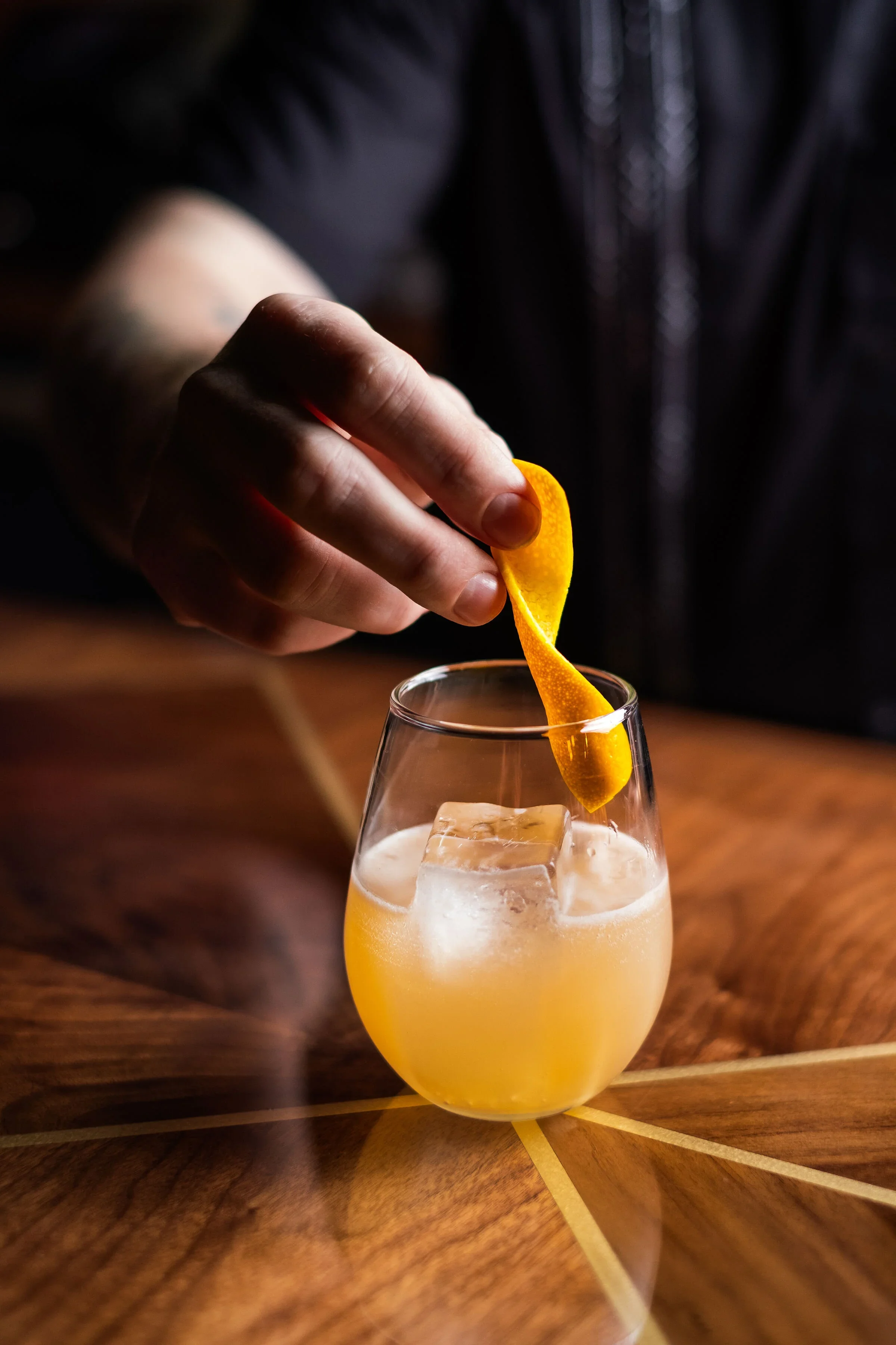 A hand placing a lemon twist into a glass of light yellow cocktail with ice cubes on a wooden table.