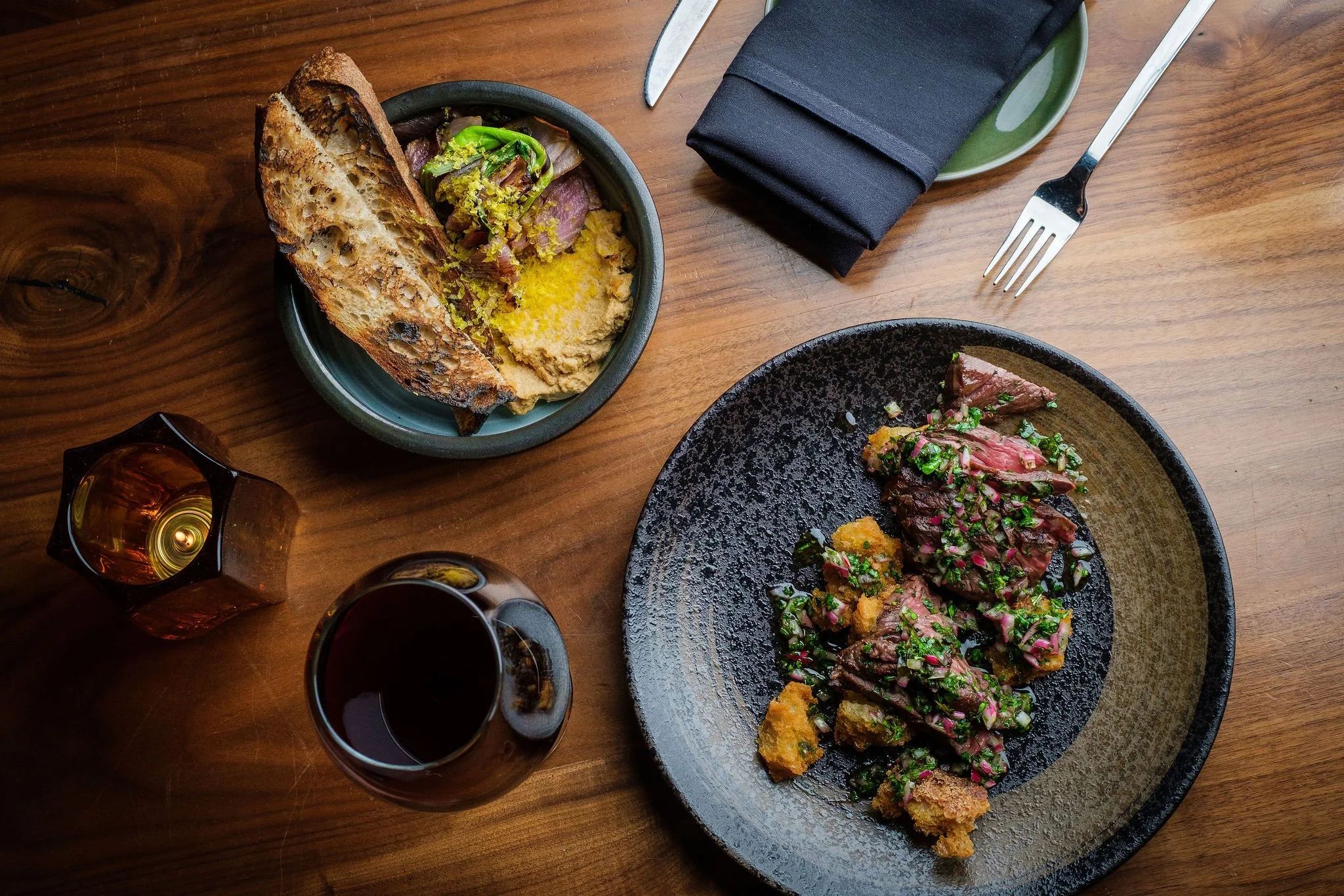 A wooden table set with a black plate of sliced beef topped with herbs and sauce, a bowl of bread with spreads and a small salad, a glass of red wine, a small candle, cutlery, and a black napkin.
