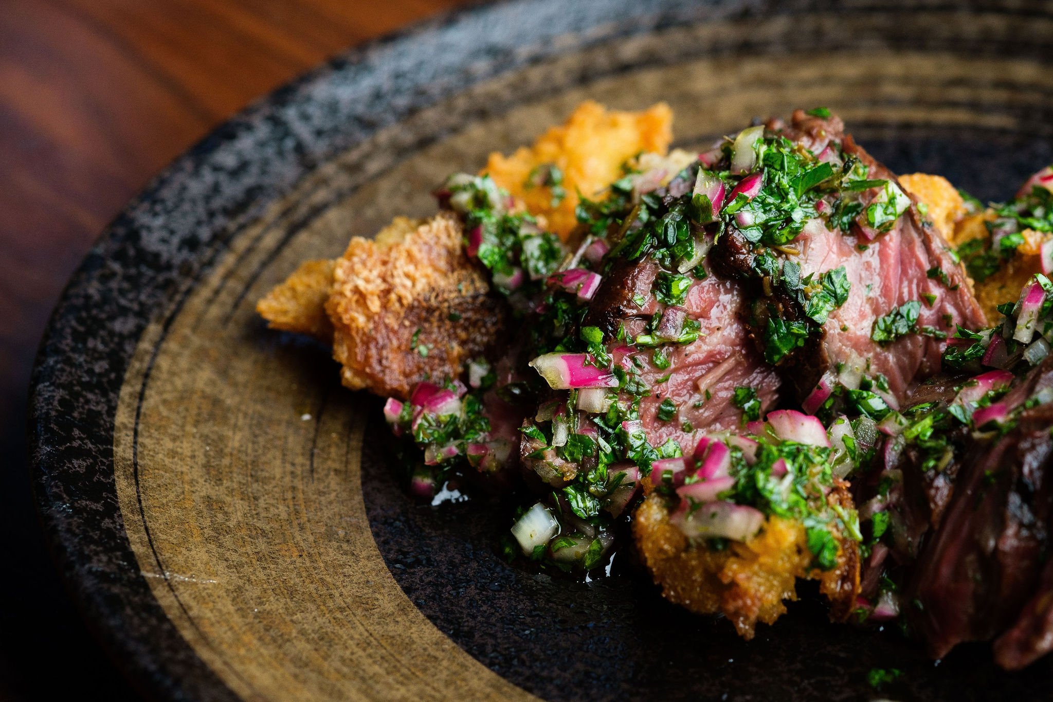 Close-up of a plate with a medium-rare cooked piece of beef topped with a green and pink chopped herb and onion mixture, accompanied by crispy fried food items on a dark wooden plate.