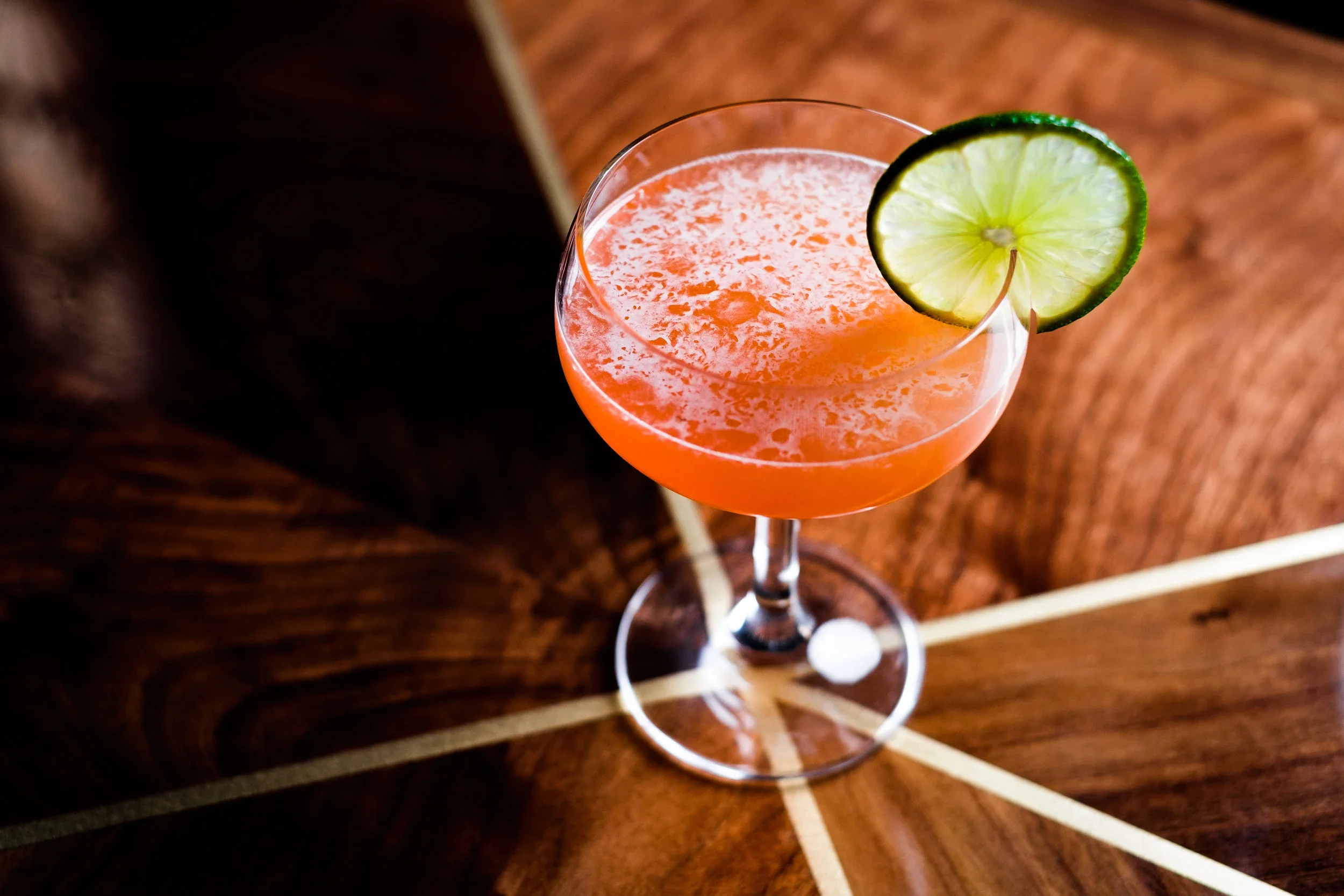 A pink cocktail with condensation in a coupe glass garnished with a lime wheel, placed on a wooden surface.