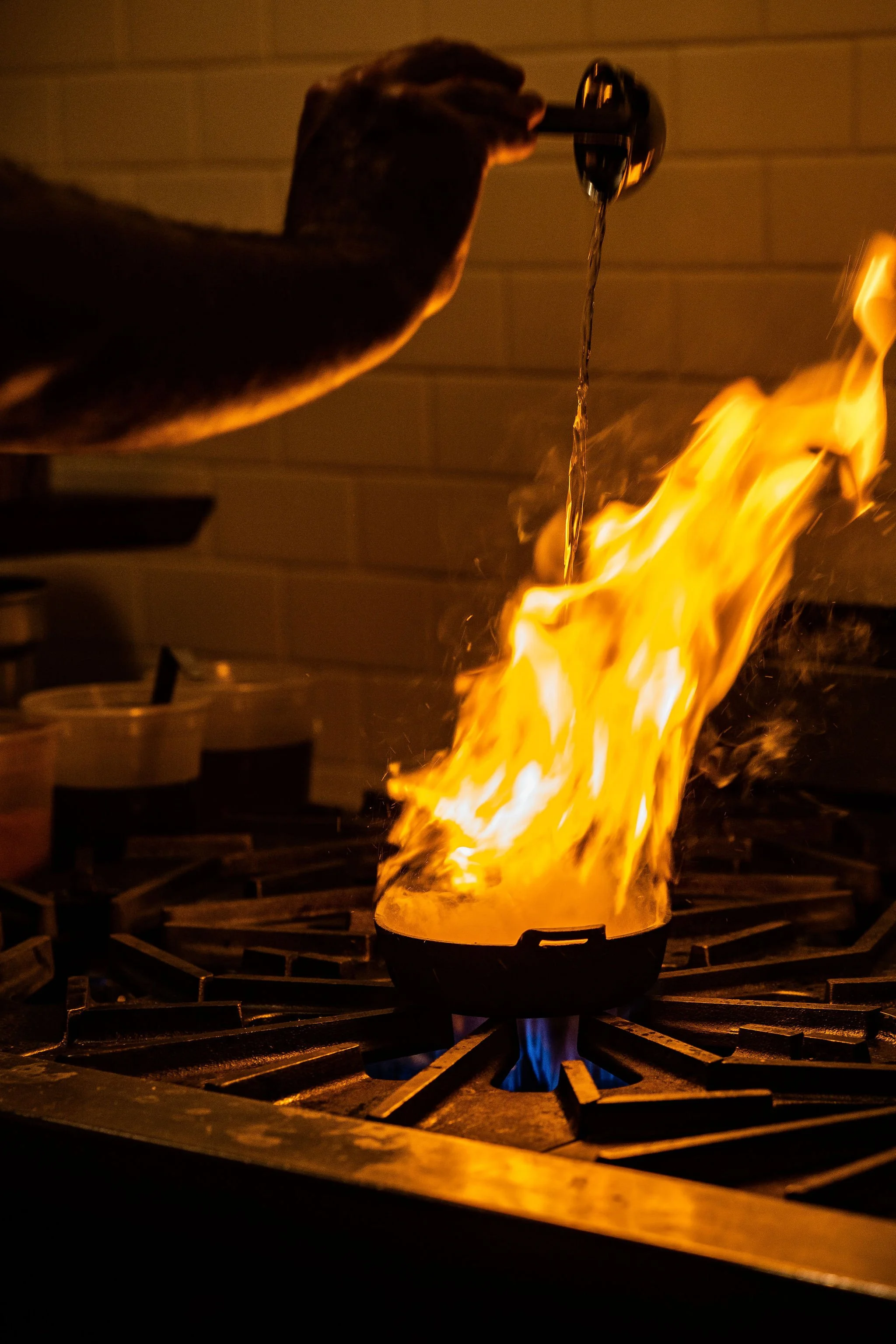 A person pouring liquid into a pan on a stove, causing a large flame and fire to erupt.