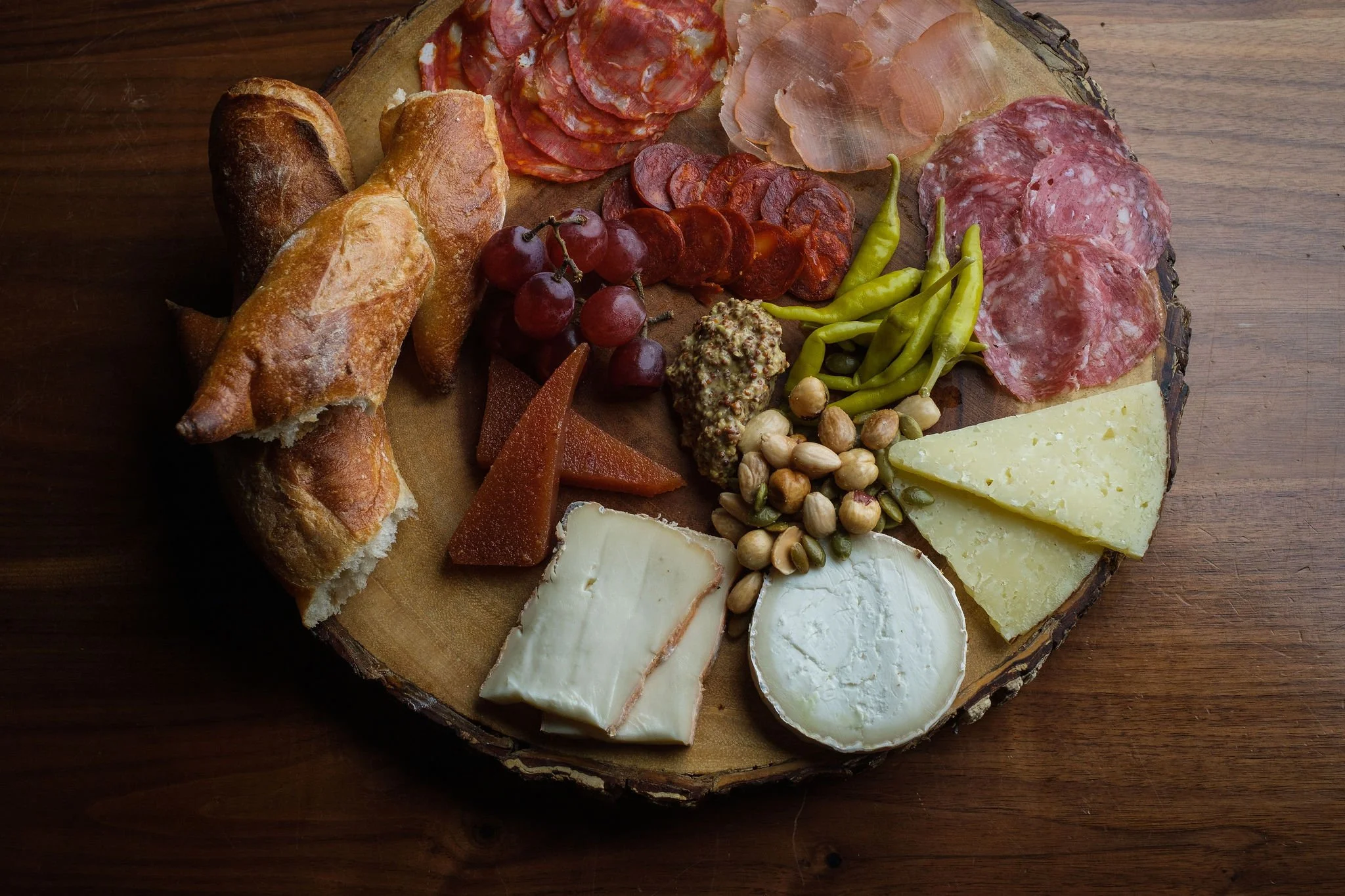 Charcuterie board with bread, various cheeses, sliced meats, grapes, nuts, and vegetables on a wooden table.