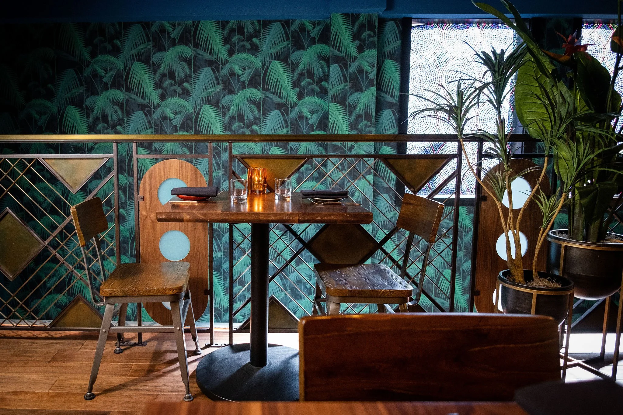 A cozy restaurant corner with a wooden table set for two, decorated with black napkins, glasses, and a candle. The background features a vibrant green tropical leaf-patterned wall, with a decorative screen or window with circular patterns and potted 