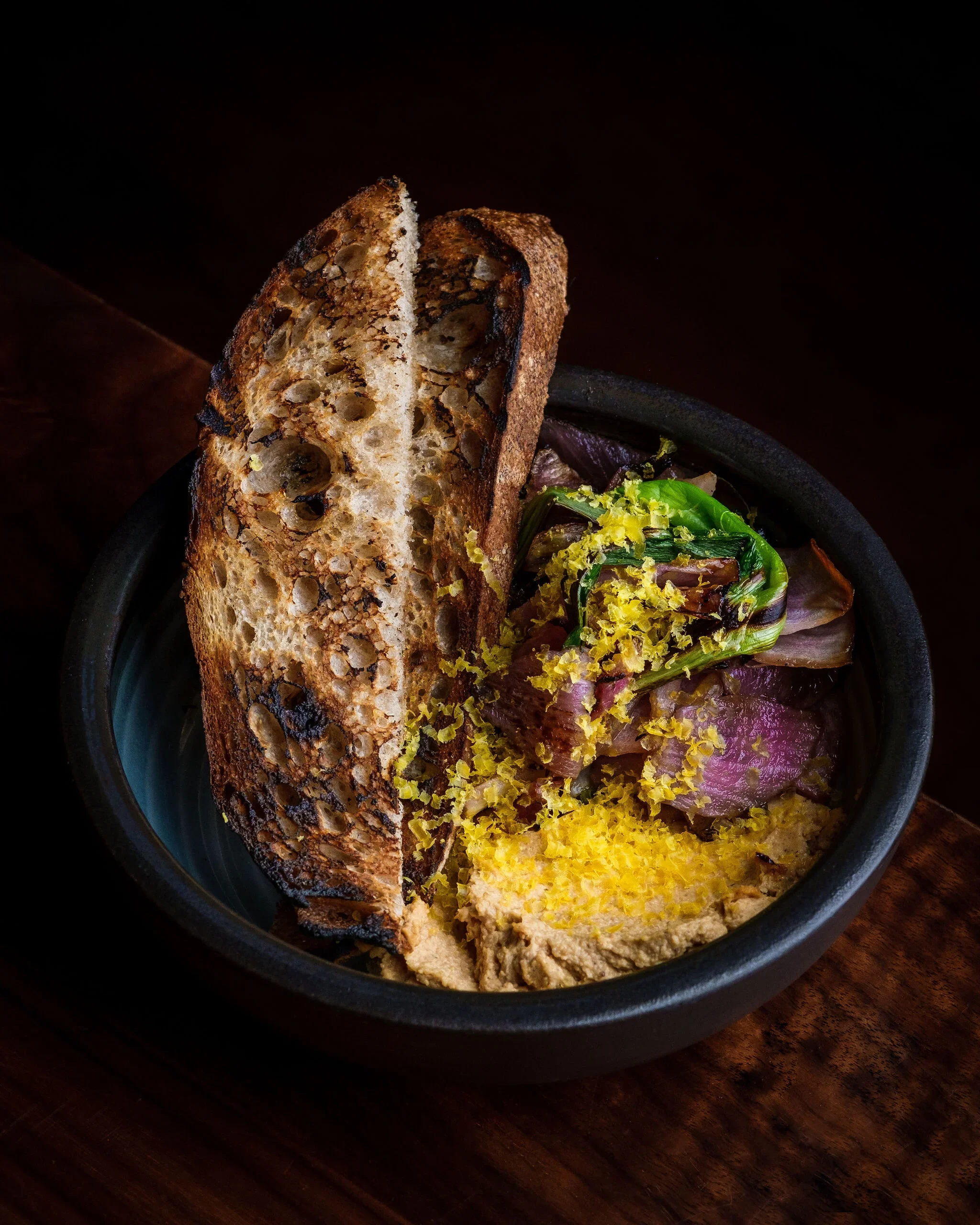A black bowl containing toasted bread, roasted vegetables, and hummus on a wooden surface.