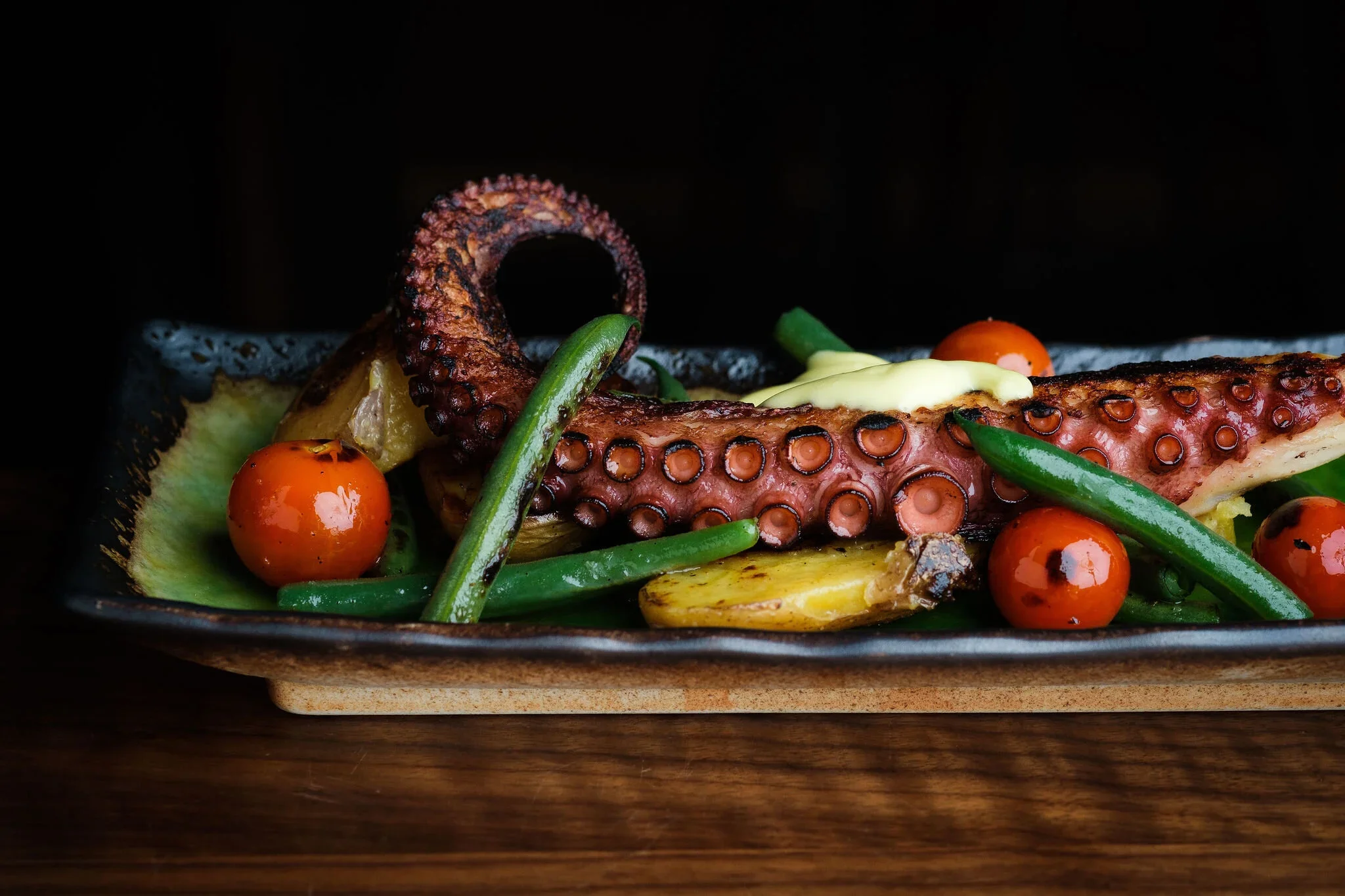 Grilled octopus tentacle with cherry tomatoes, green beans, and yellow potatoes on a black rectangular plate.
