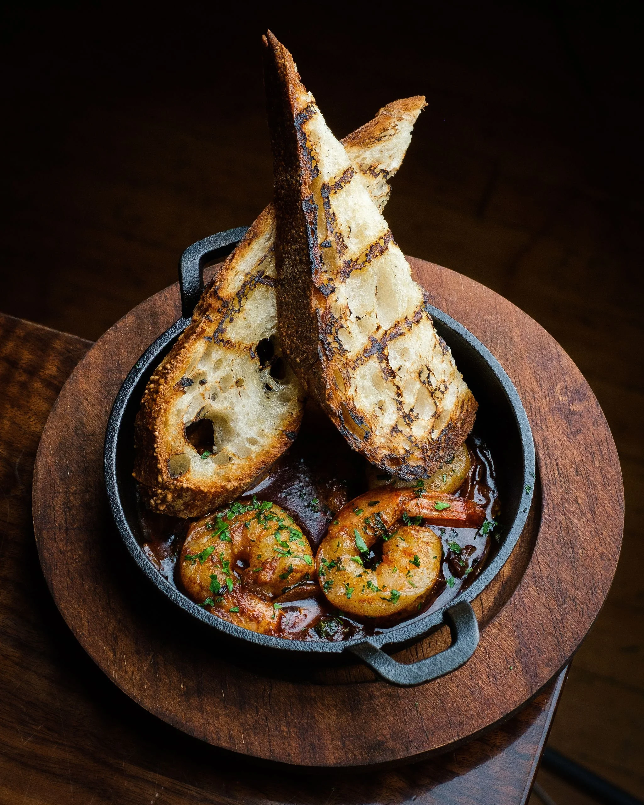 A cast iron skillet containing shrimp, vegetables, and sauce, topped with toasted bread slices, on a wooden serving board.