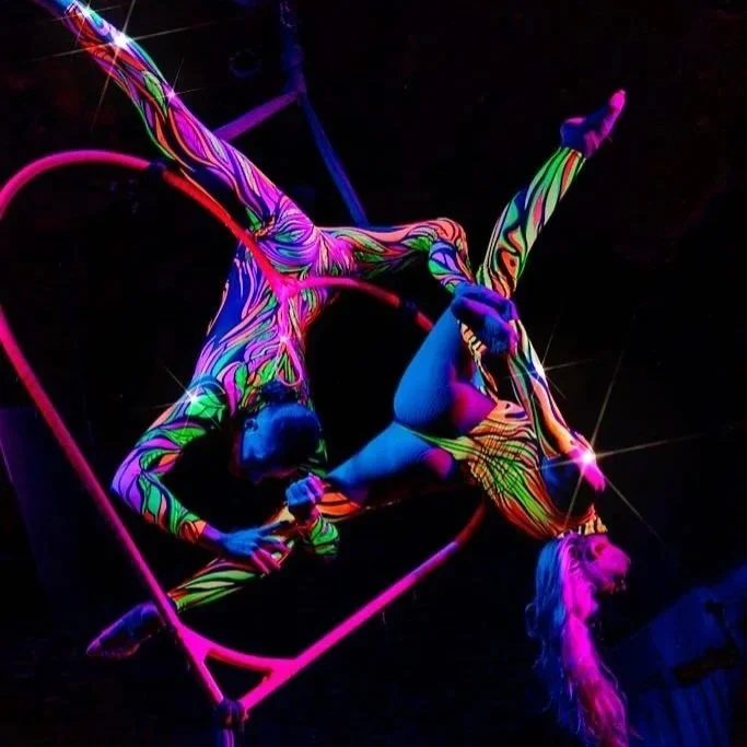 Two performers from 200 Channels in neon body paint performing acrobatics on a pink trapeze under black lights.