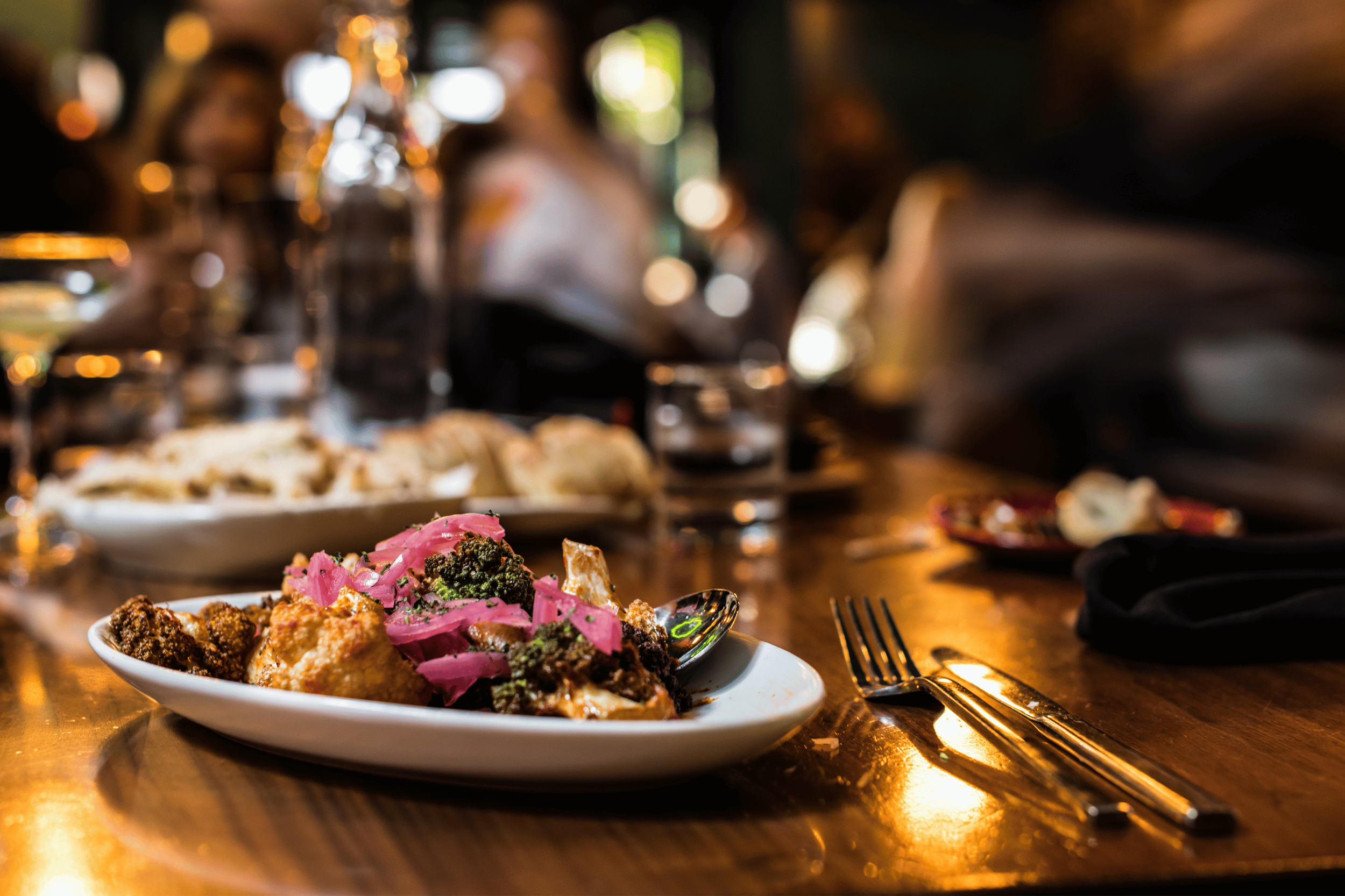 A close-up of a plate of food on a restaurant table, with a blurry background of people dining. The dish contains fried items topped with pink pickled onions and herbs, and a spoon resting on the plate.