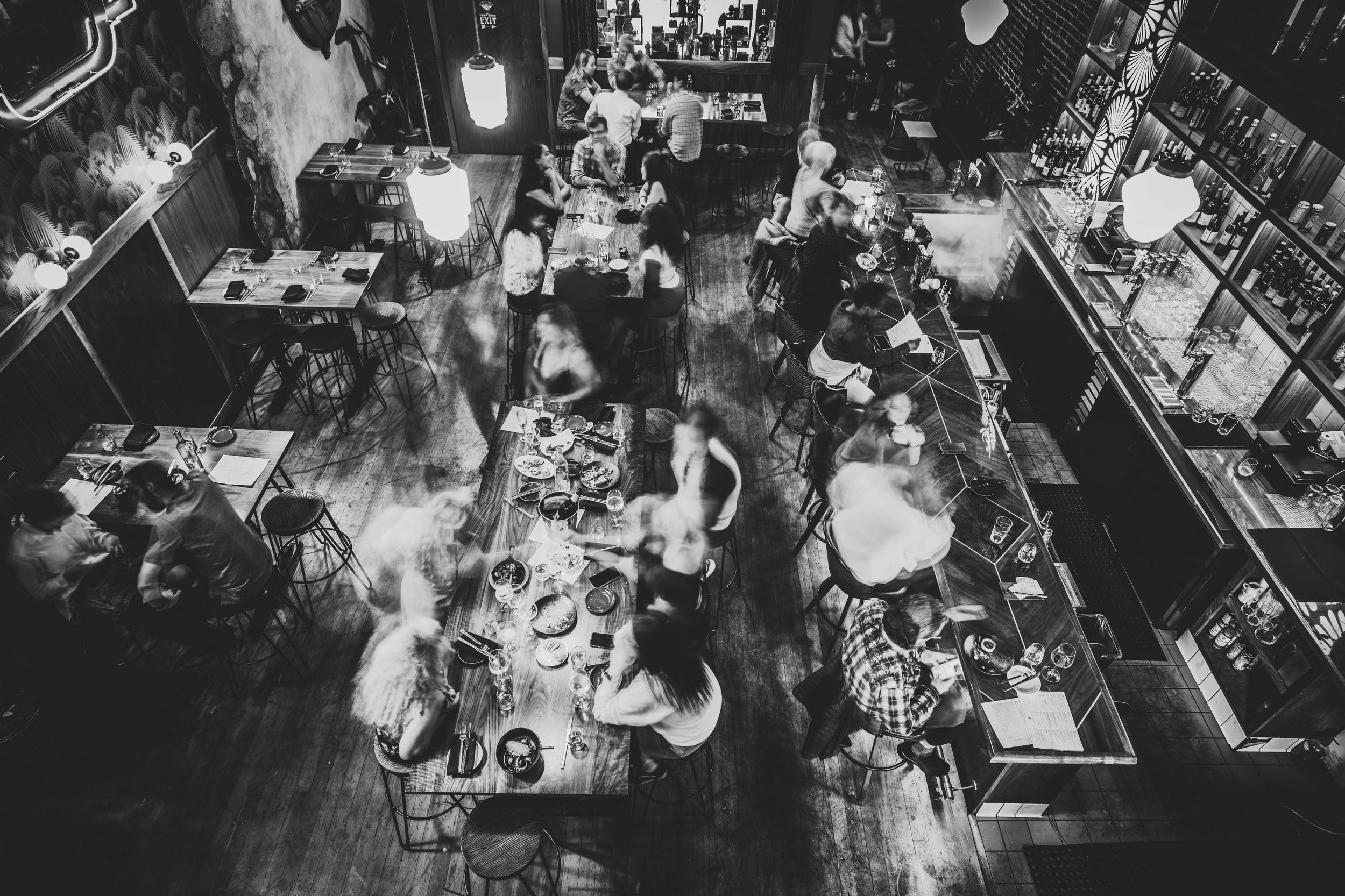 Top-down view of a busy night at The Pawn Shop restaurant and bar with patrons sitting at tables and a bar counter, black and white photo, some blurriness from motion.