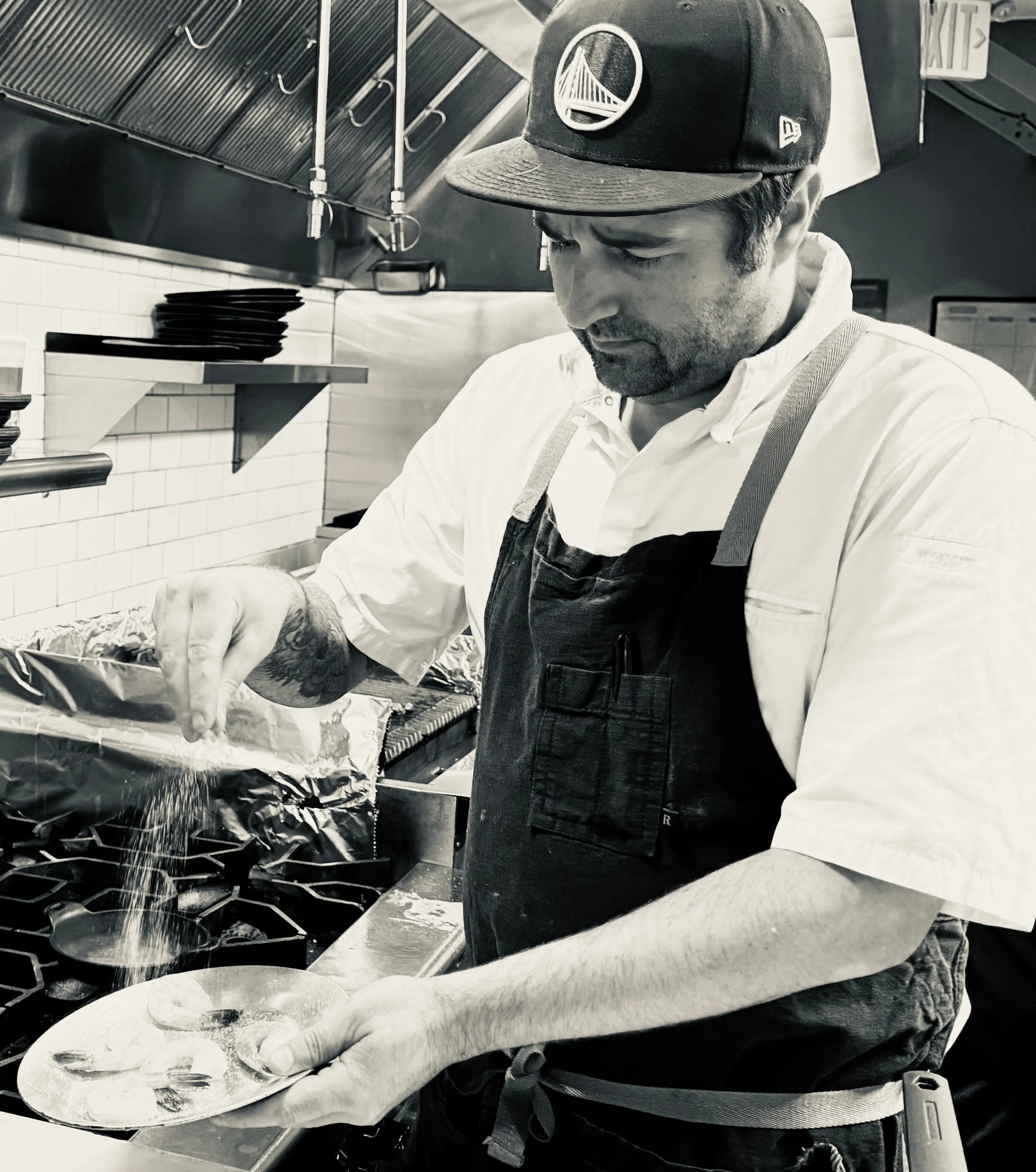 A chef sprinkling seasoning onto a dish in a professional kitchen.