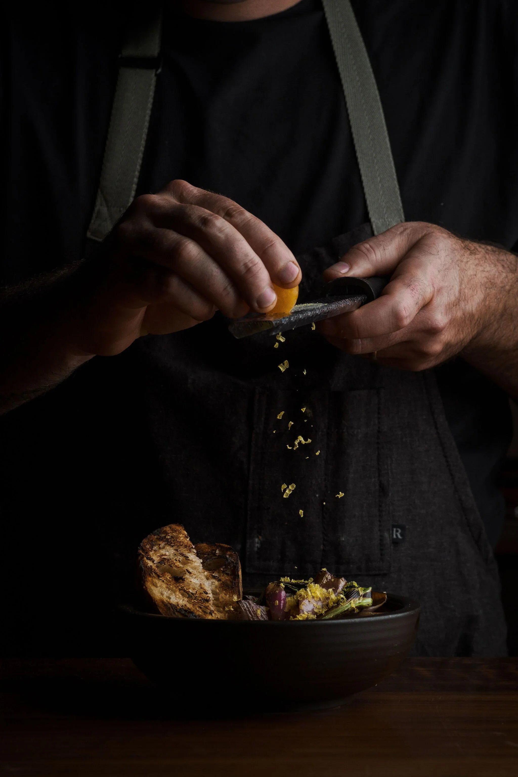 Person grating lemon zest over a bowl of salad, wearing a black shirt and apron.