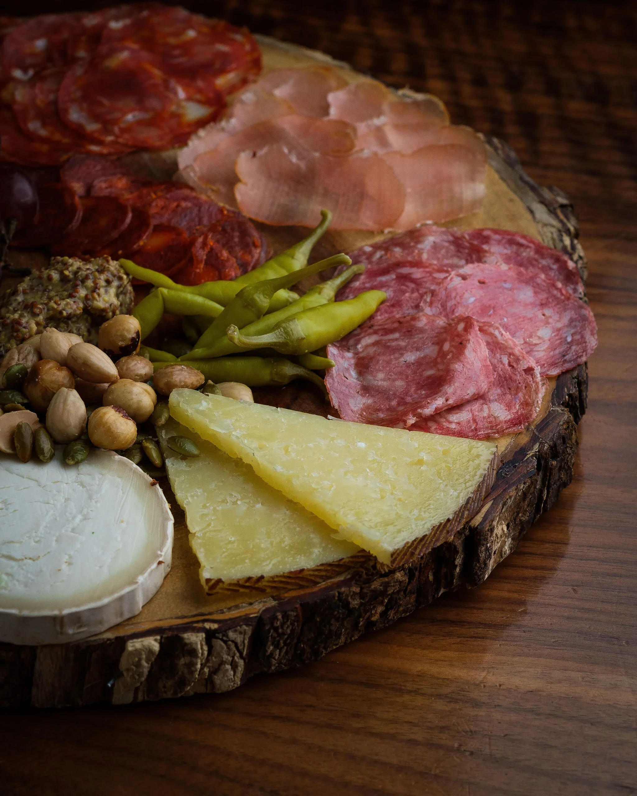Assorted cured meats, cheeses, green chili peppers, nuts, and bread on a wooden serving board.