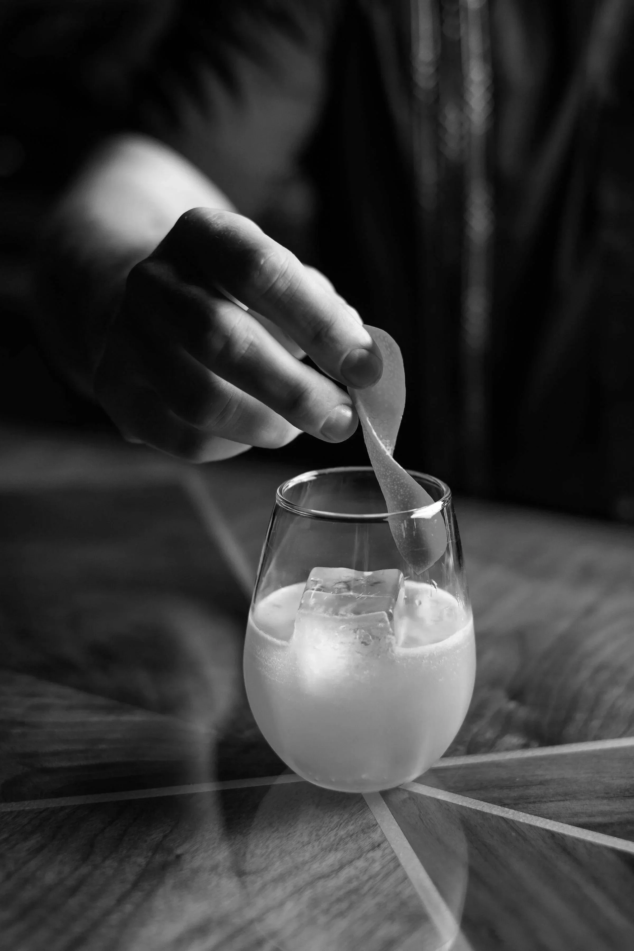A person placing a spoon into a cocktail glass on the bar at The Pawn Shop filled with a beverage and ice cubes, taken in black and white.