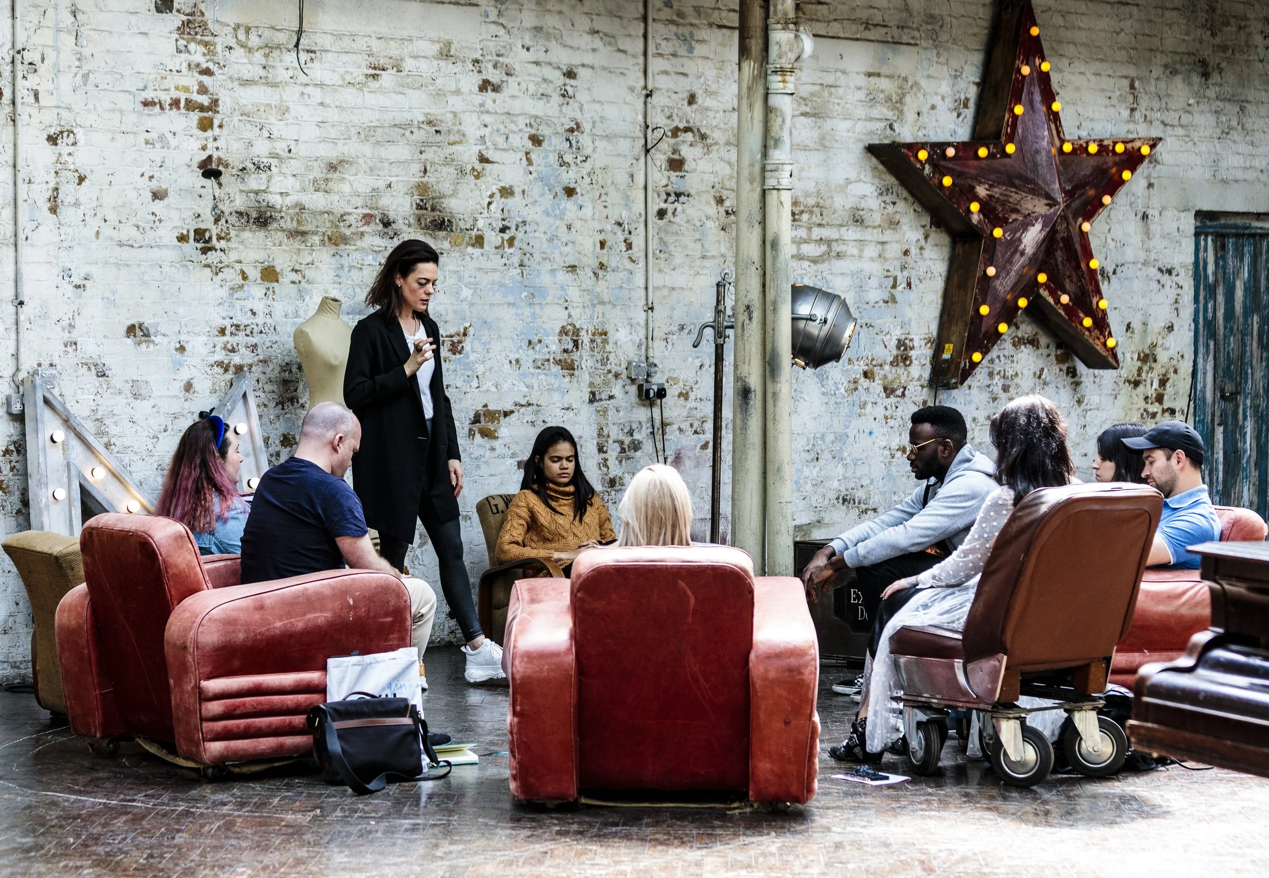 A group of people sitting in a circle on vintage chairs inside an industrial-style room with a large lit star decoration on the wall behind them. The room's decor includes exposed brick, a mannequin, and stage lighting. The setting has a casual and creative atmosphere.