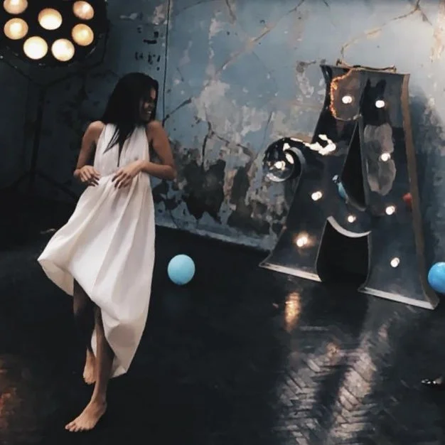 Woman in white dress dancing near illuminated letter A with distressed wall background.