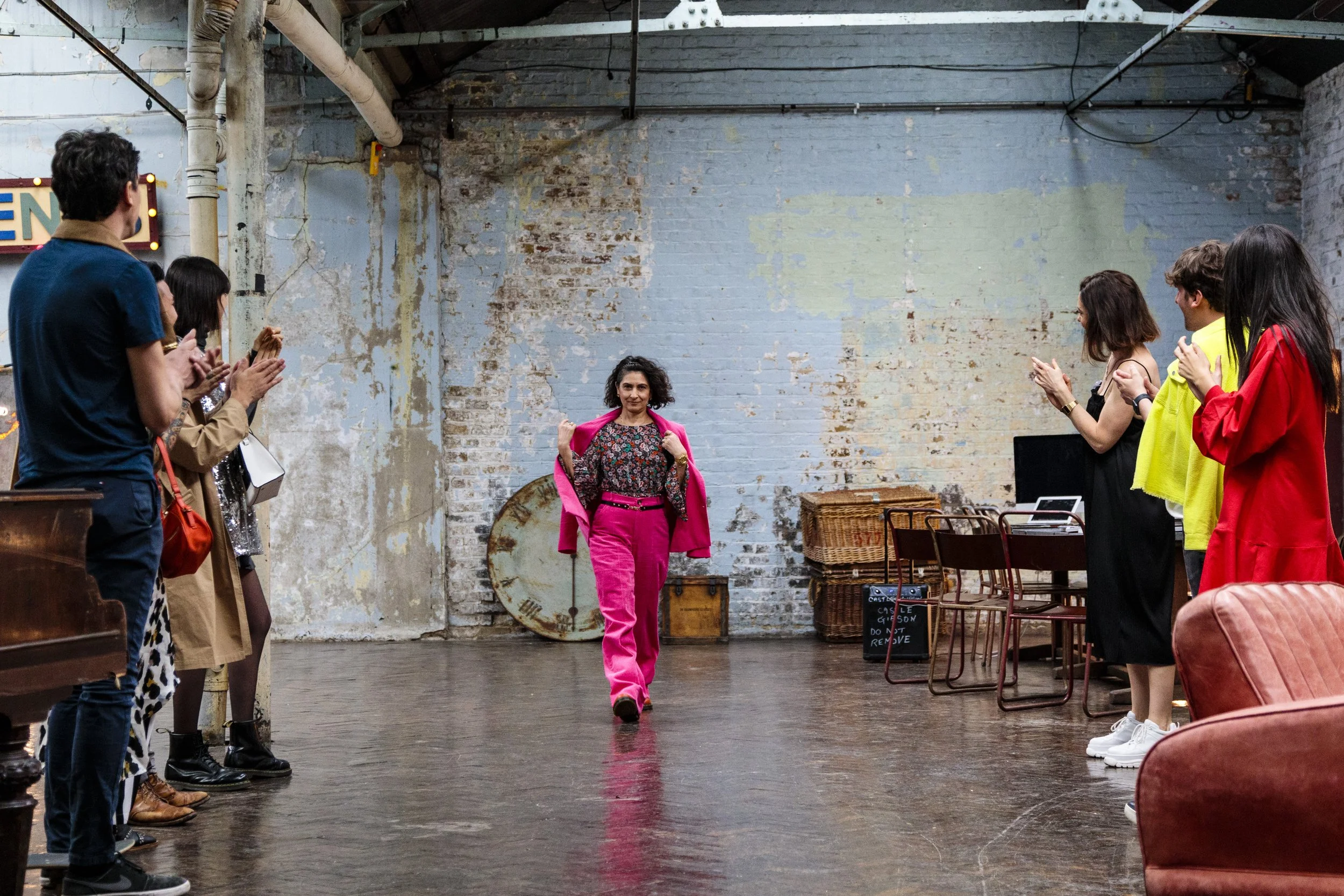 A person in a hot pink outfit walking confidently in an industrial-style room, flanked by people clapping along the sides.