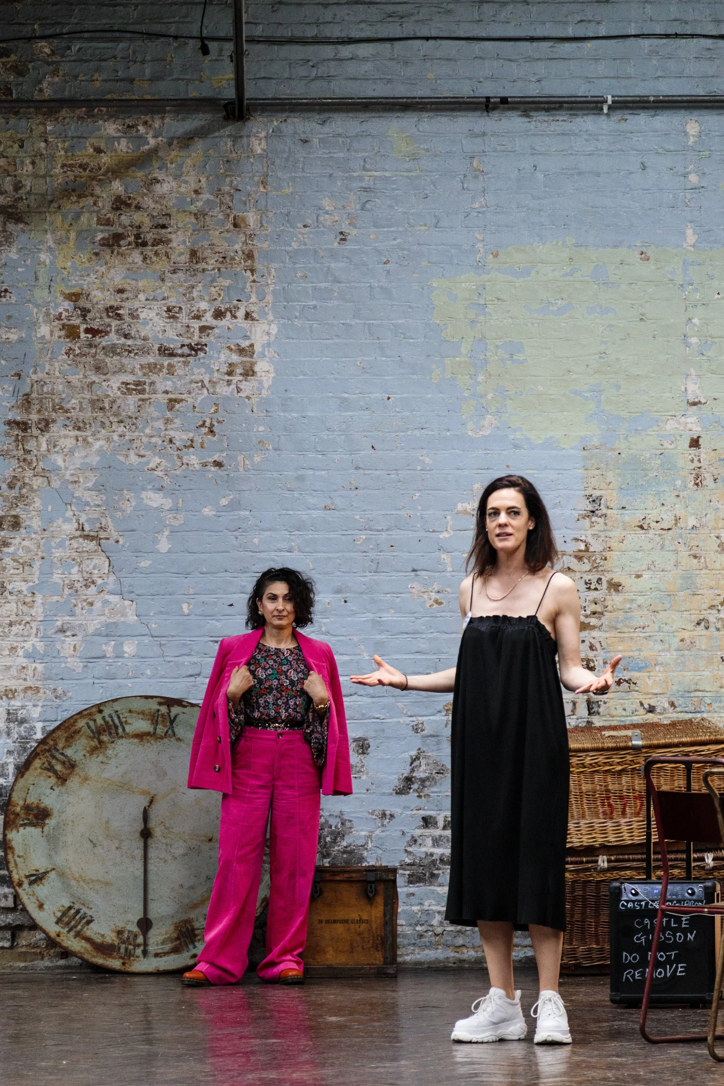 Two people in a rustic room; one in a pink suit and floral top, one in a black dress with white sneakers, standing near a wall with peeling paint and vintage clocks.