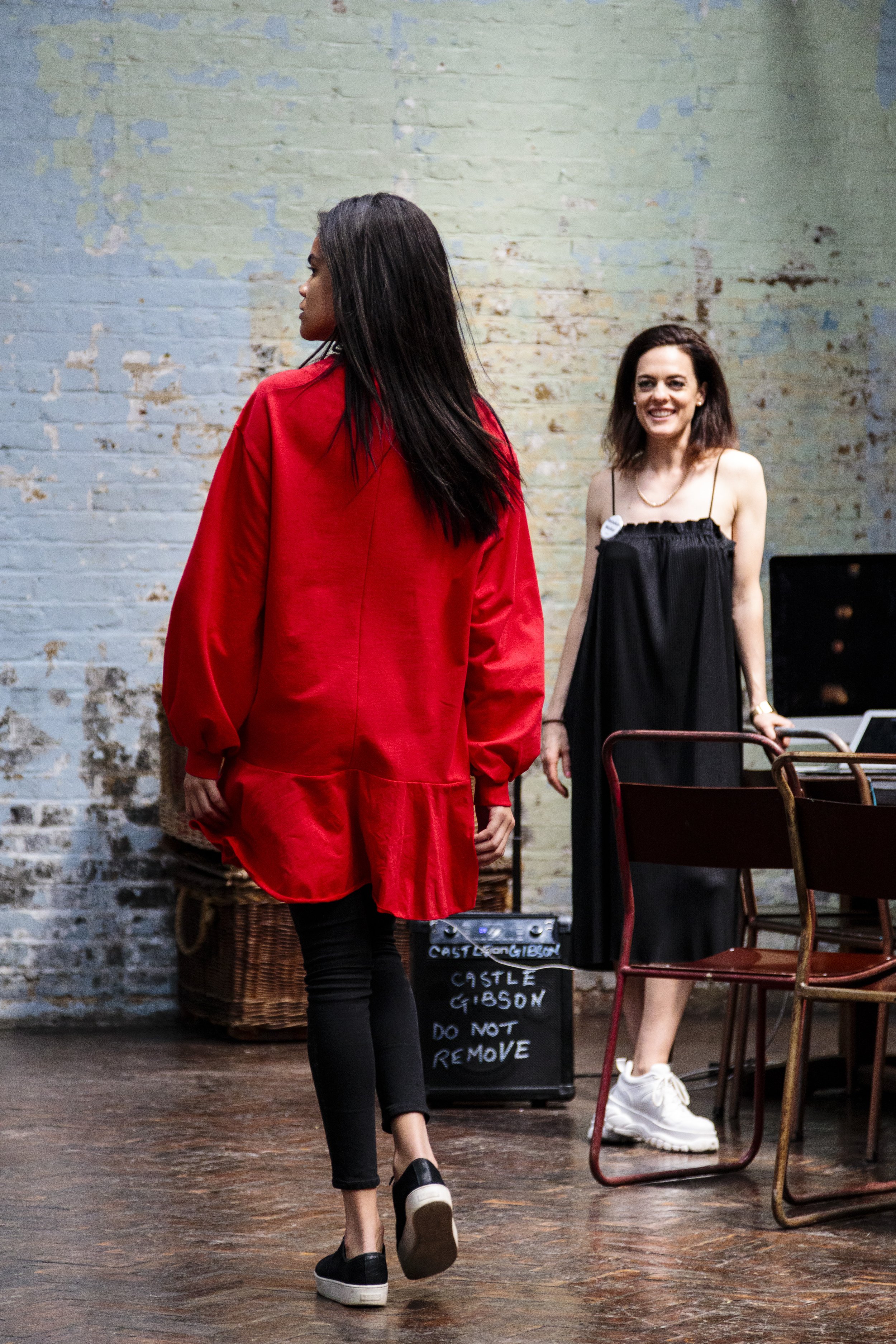 Two women in a room, one wearing a red top and black pants, walking, and the other in a black dress, smiling. The background shows a brick wall and some chairs.