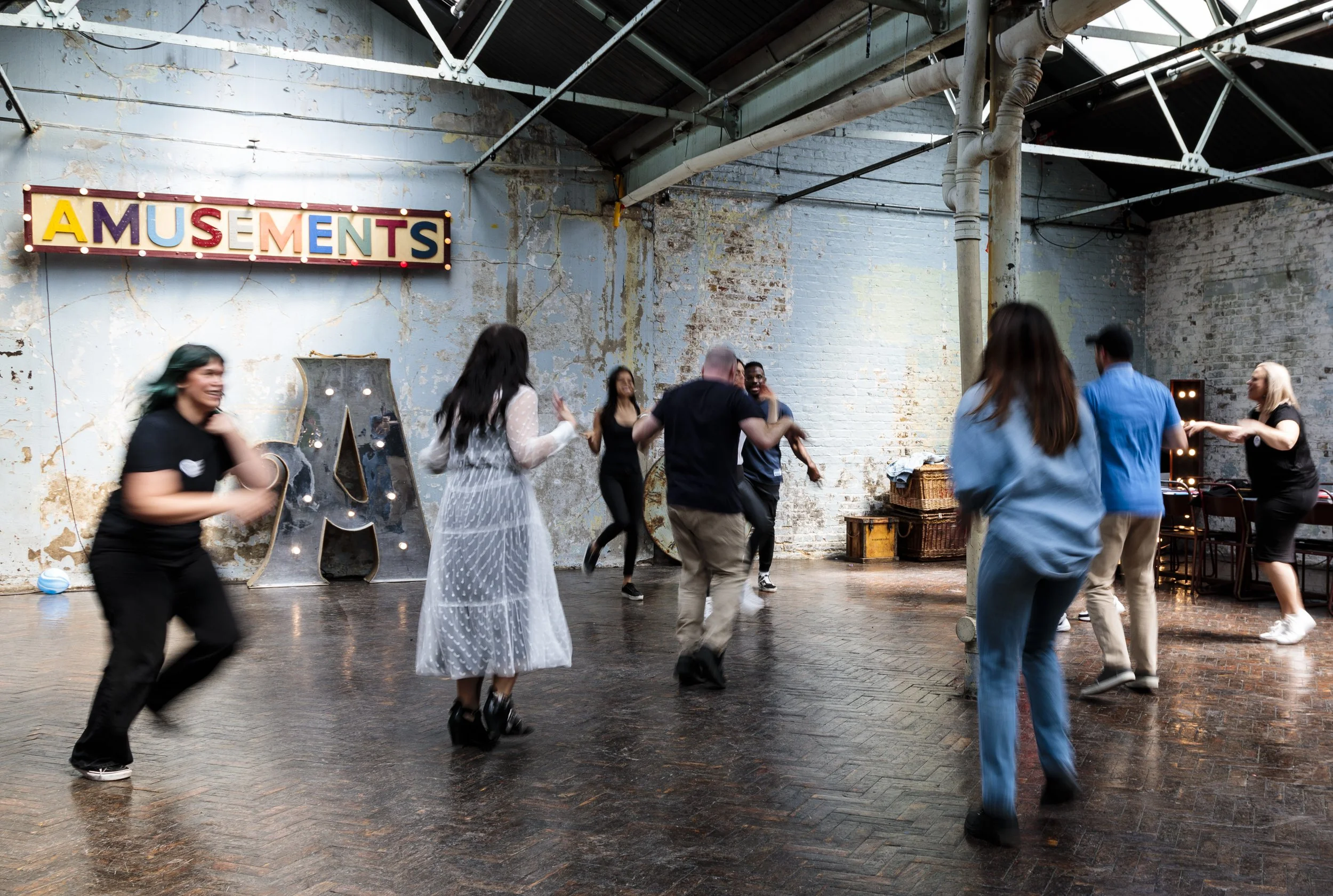 Group of people dancing energetically in an industrial-style room with exposed brick. A sign reading 'Amusements' is on the wall. A large letter 'A' with lights is on the floor.