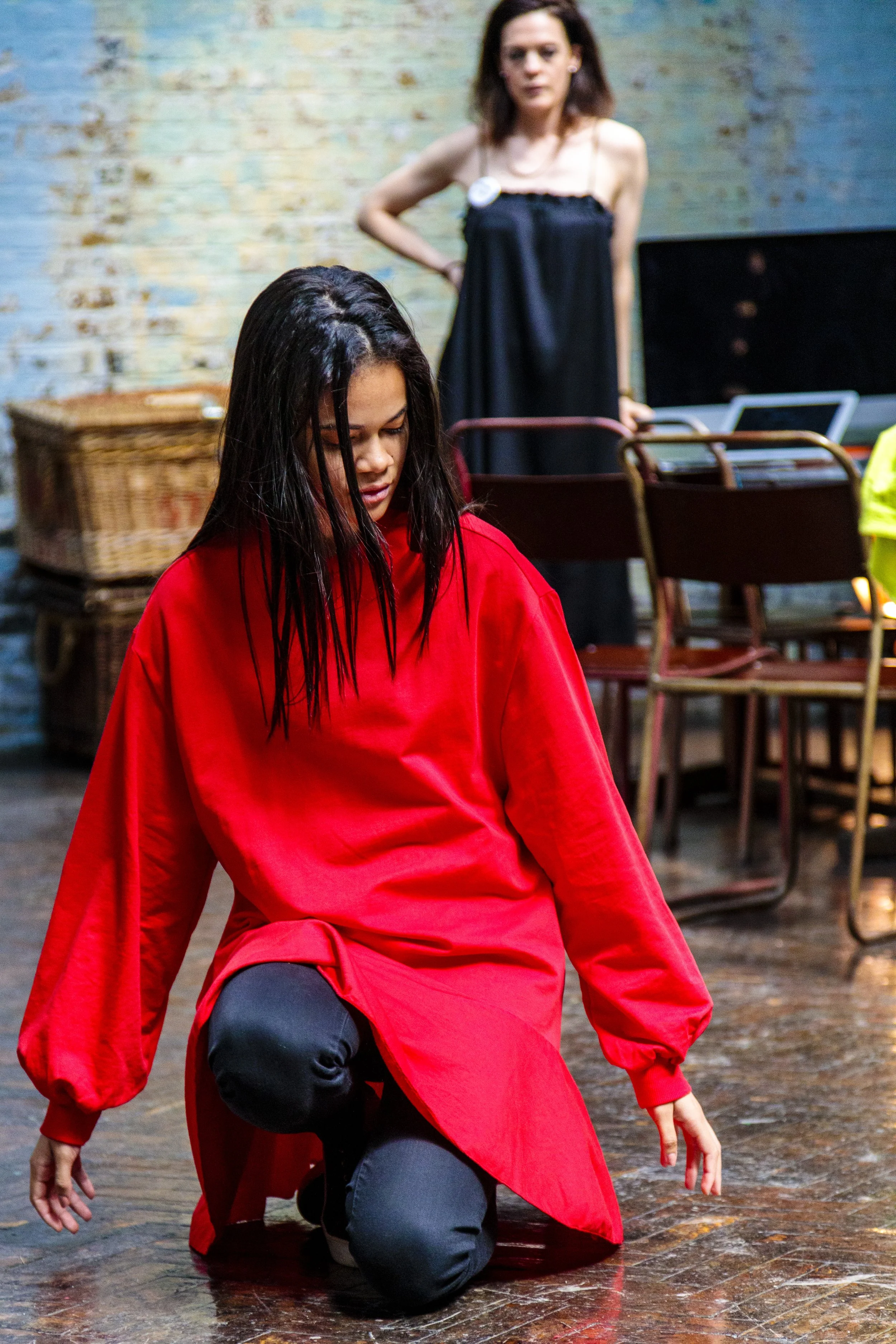 Two women in a room with brick walls, one kneeling in a red dress, the other standing in a black dress with hands on hips, surrounded by chairs and baskets.