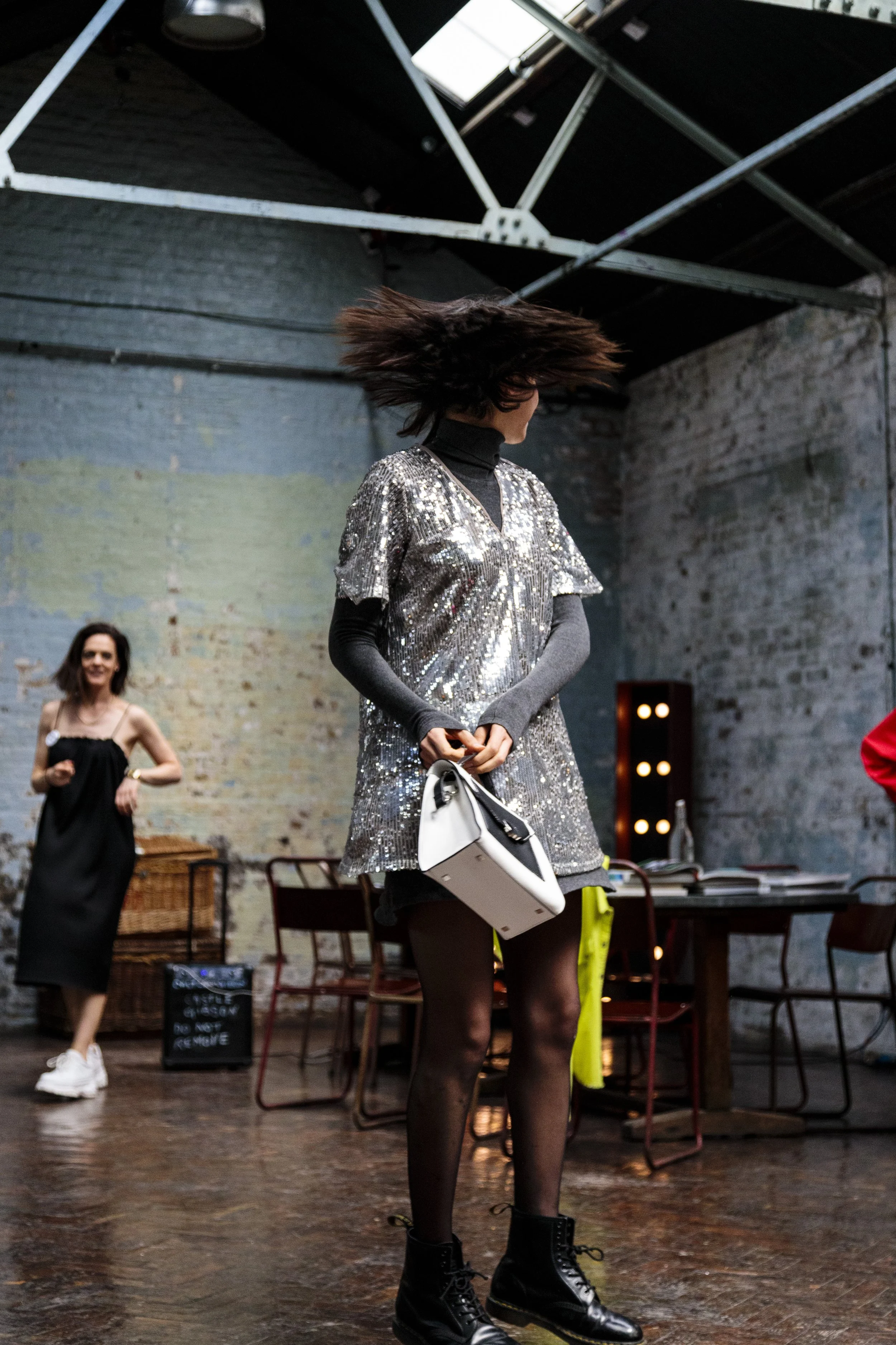 Woman in a sparkly silver dress holding a white handbag, with another woman in a black dress in the background, in an urban industrial setting.