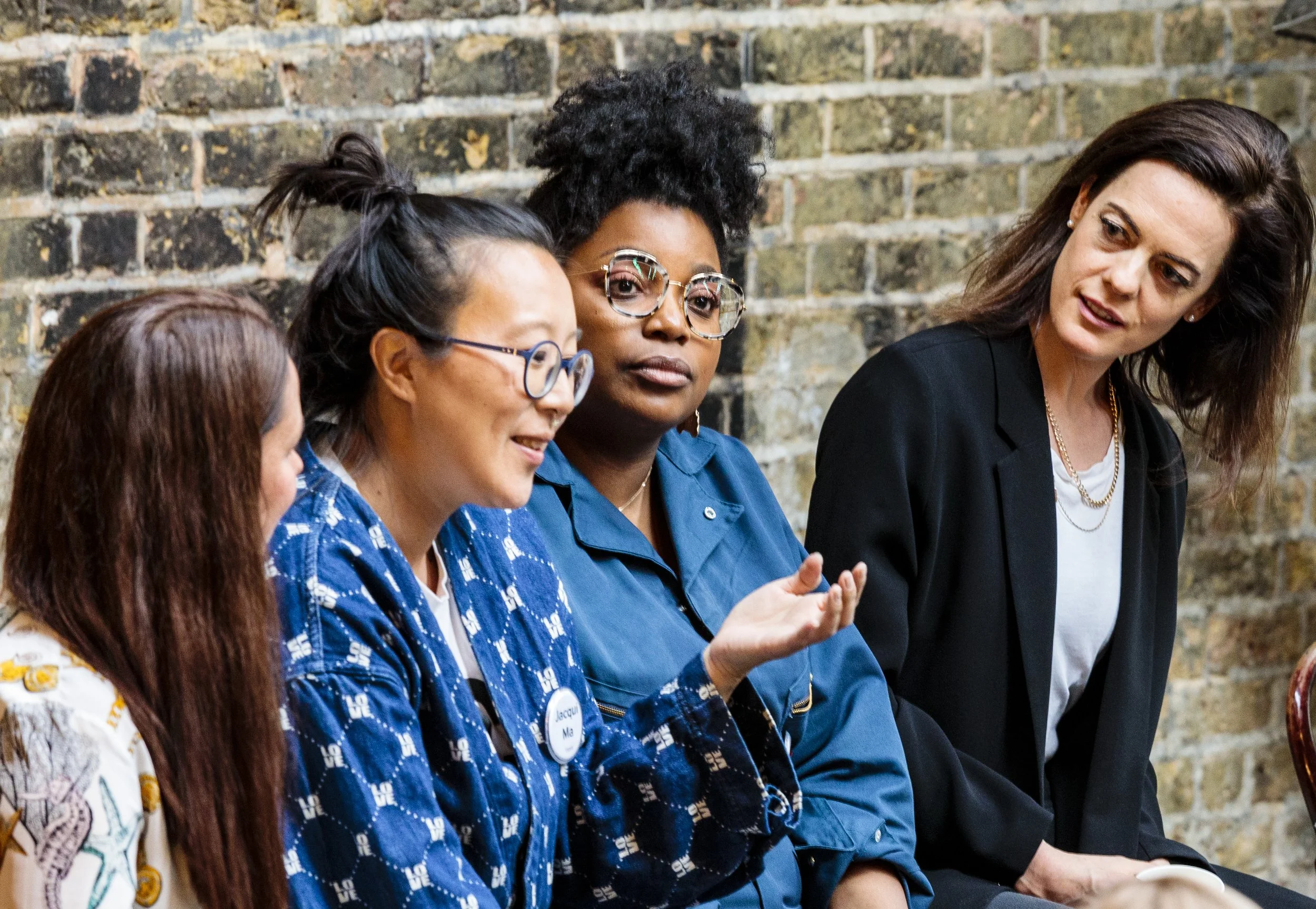 Four women sitting and engaged in a discussion against a brick wall backdrop. One woman gestures while speaking, and two others listen attentively.