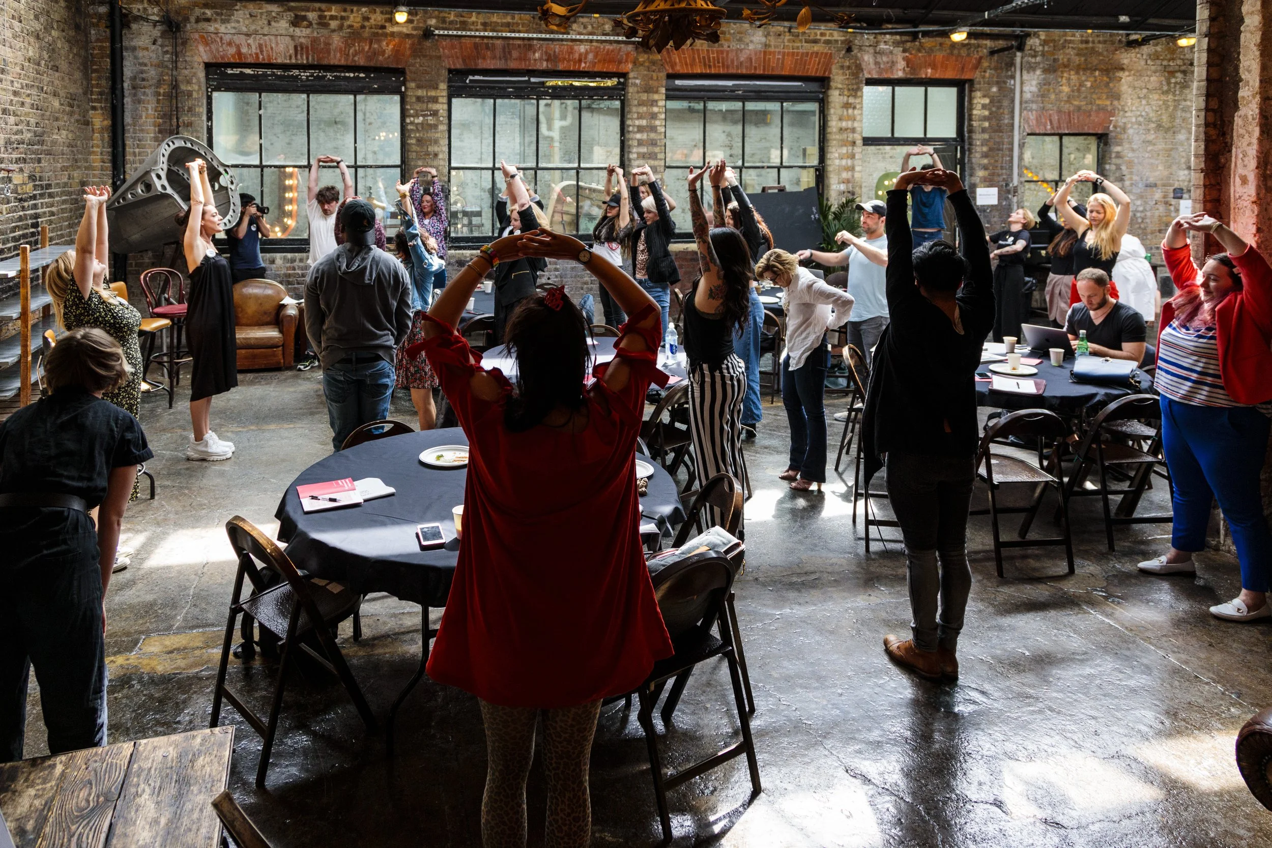 People in a workshop doing group stretching exercises in a rustic, industrial-style room with brick walls and large windows.