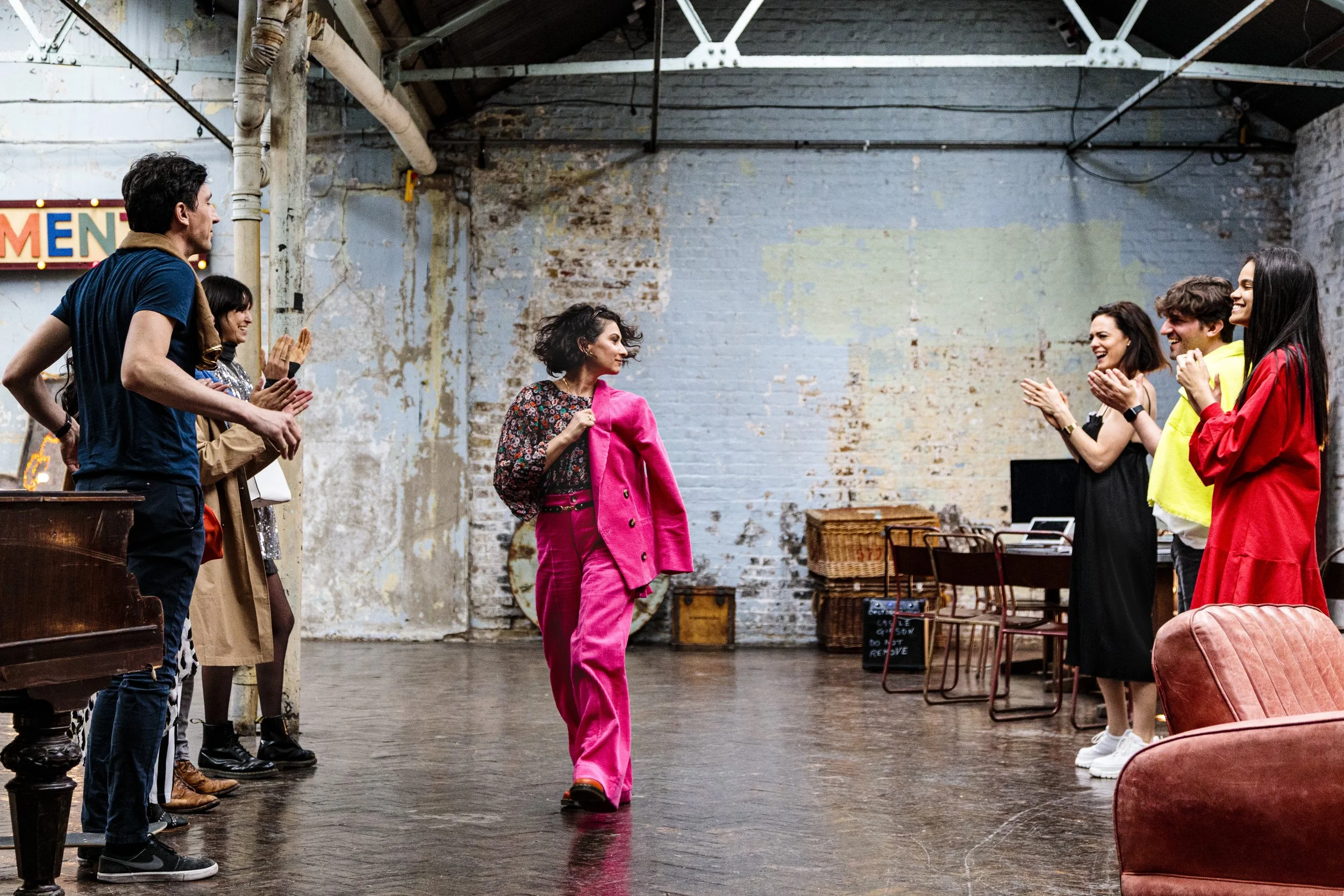 A person in a bright pink suit walks confidently in an industrial-style room, while a group of people applaud. The room has exposed brick walls and pipework, wooden floors, and assorted furniture.