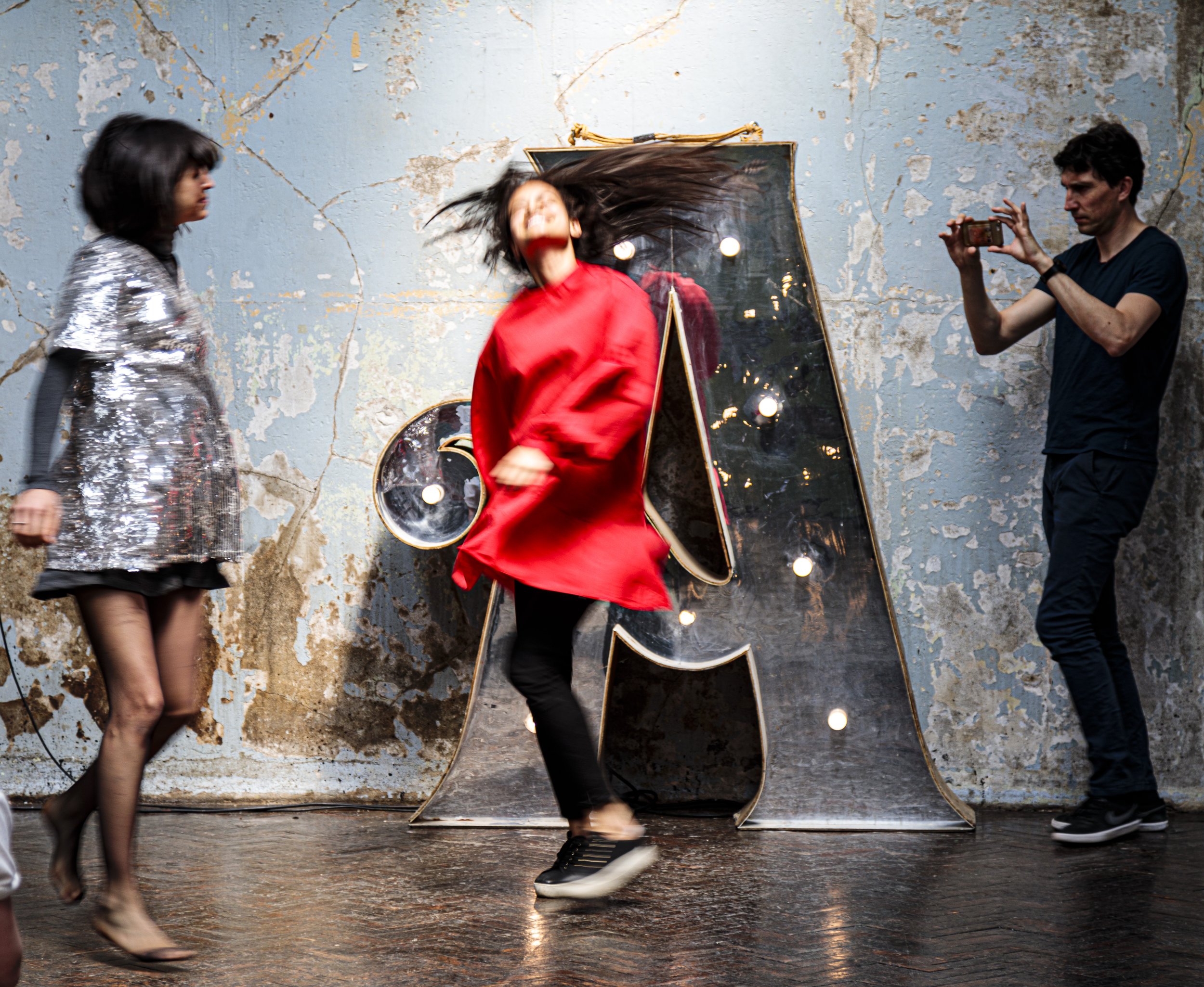 Three people in a room with a large illuminated letter "A" against a textured wall. A woman in a shiny silver dress and another in a red outfit are moving, while a man takes a photo with his phone.