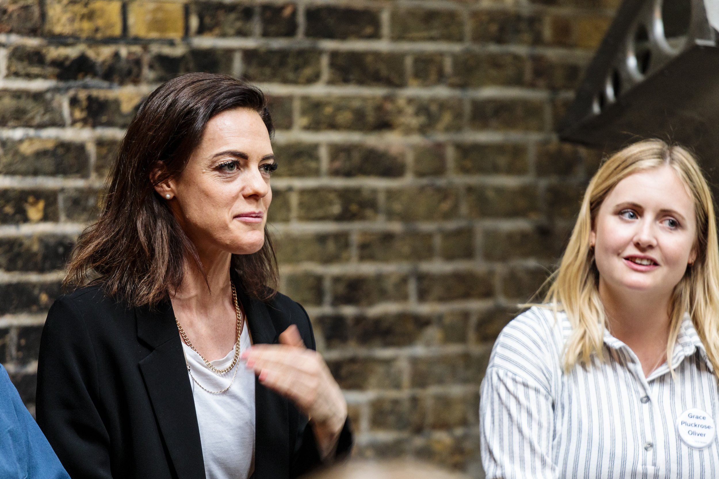Two women sitting and talking, with a brick wall background. One woman has dark hair and is wearing a black blazer, while the other has blonde hair and is wearing a striped shirt with a name tag.