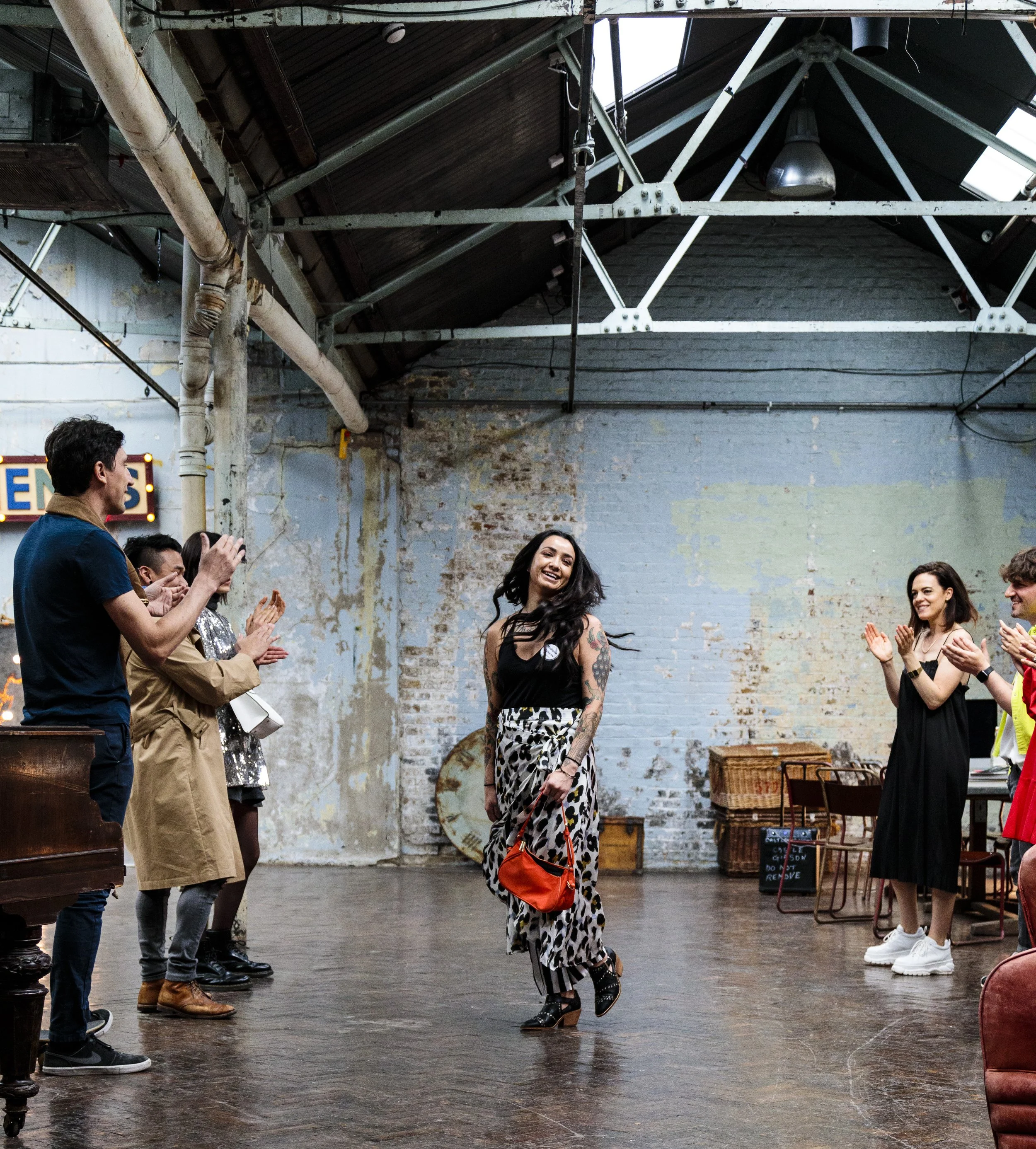 A group of people in a warehouse-style space with high ceilings, watching a woman in a patterned skirt and holding a small bag. Others are clapping and smiling, creating a lively and supportive atmosphere.