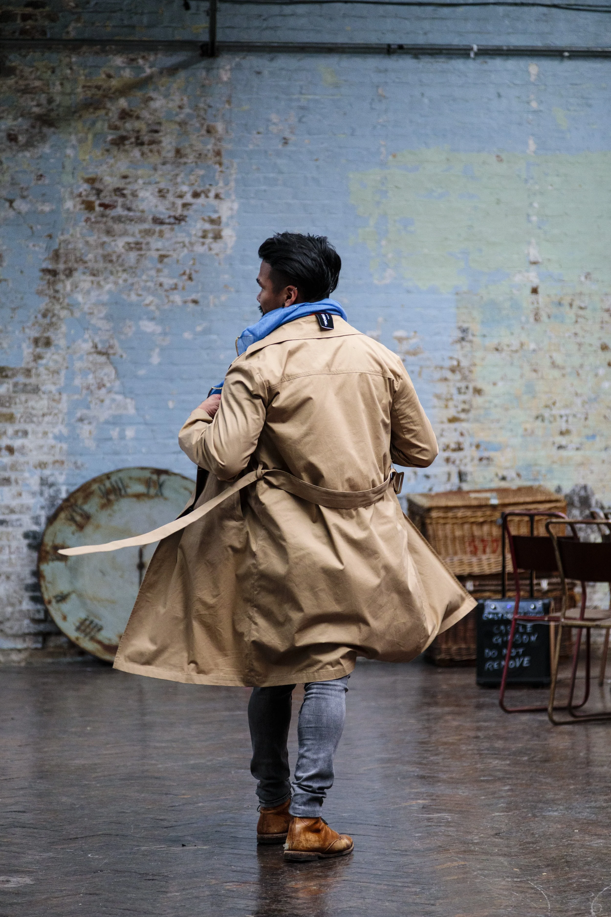 Man in a tan trench coat and blue scarf walking indoors with a rustic background.