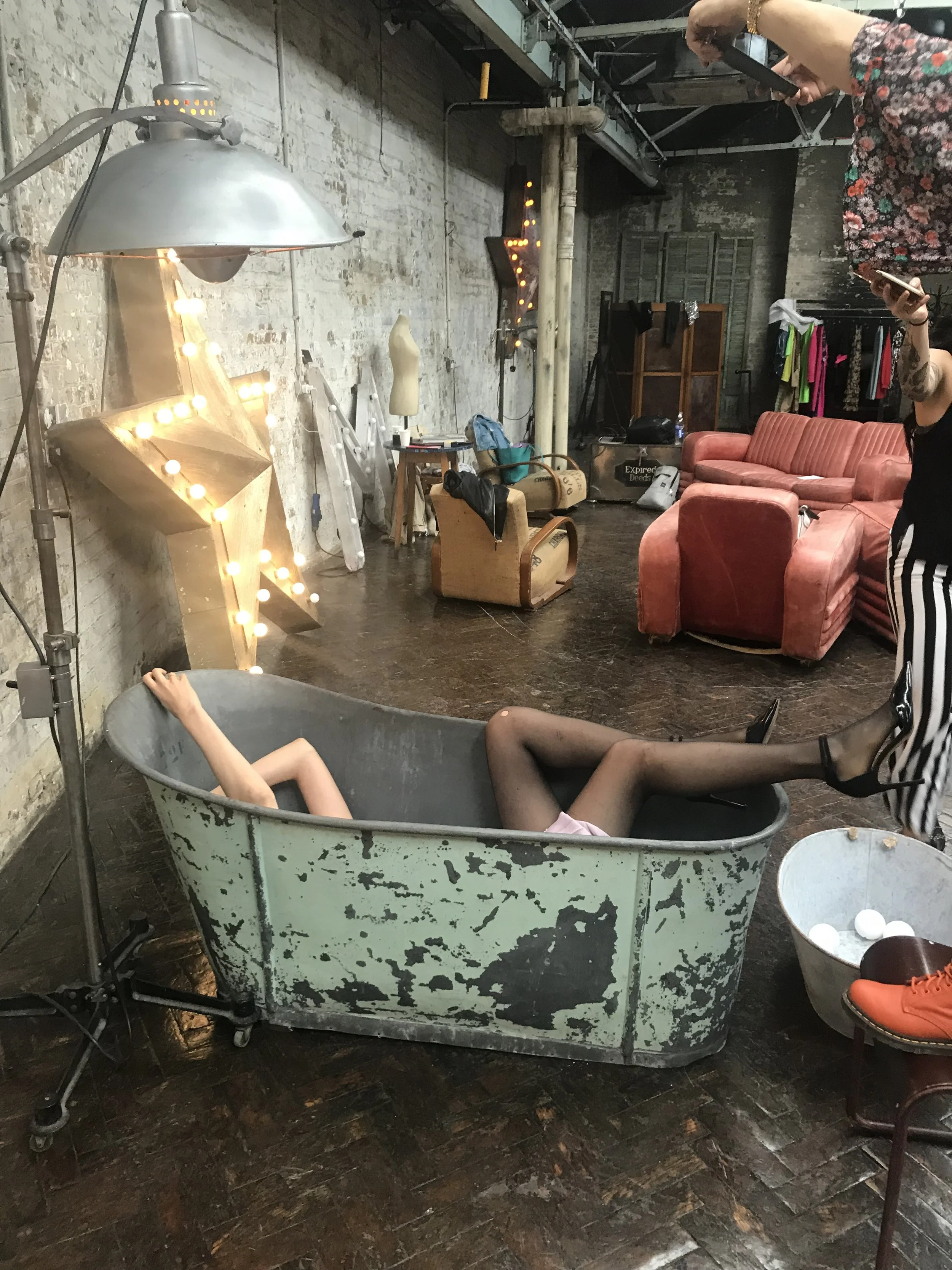 Industrial-style room with a vintage bathtub containing two mannequin legs, a metal lamp, a large lit star, and various vintage furniture and clothing items.