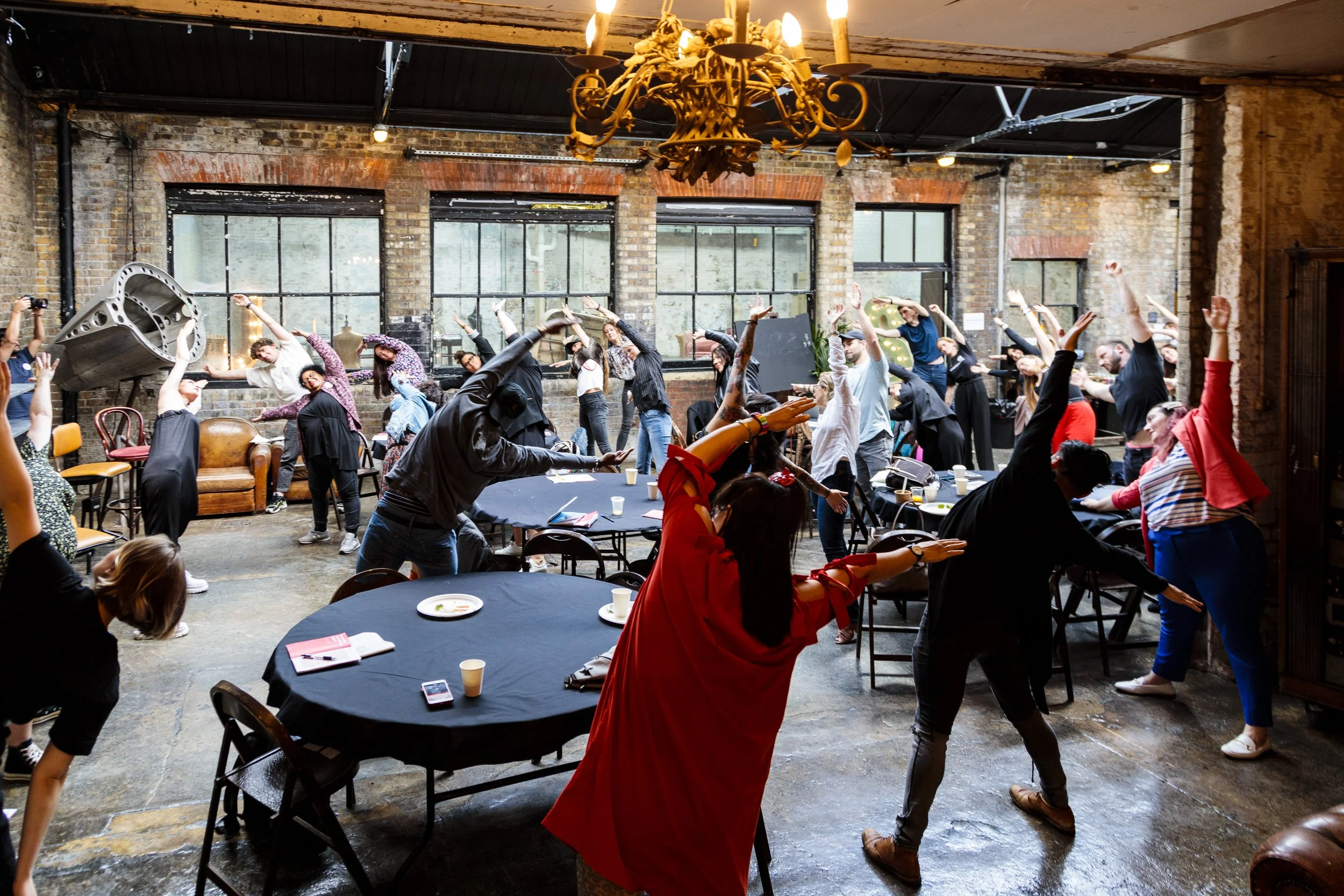 A group of people in a rustic room participating in a group stretch with tables and chairs around them.