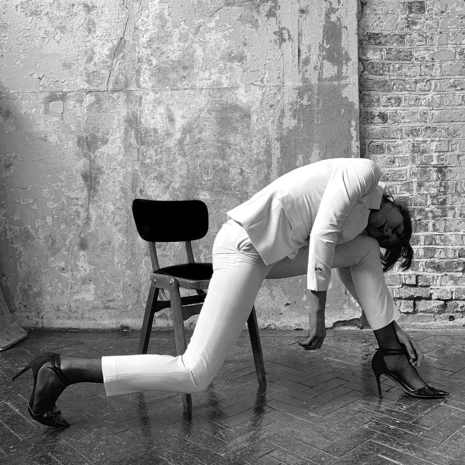 Person in an elegant pose wearing a light-colored suit and high heels, bending over next to a chair, in front of a textured industrial wall.