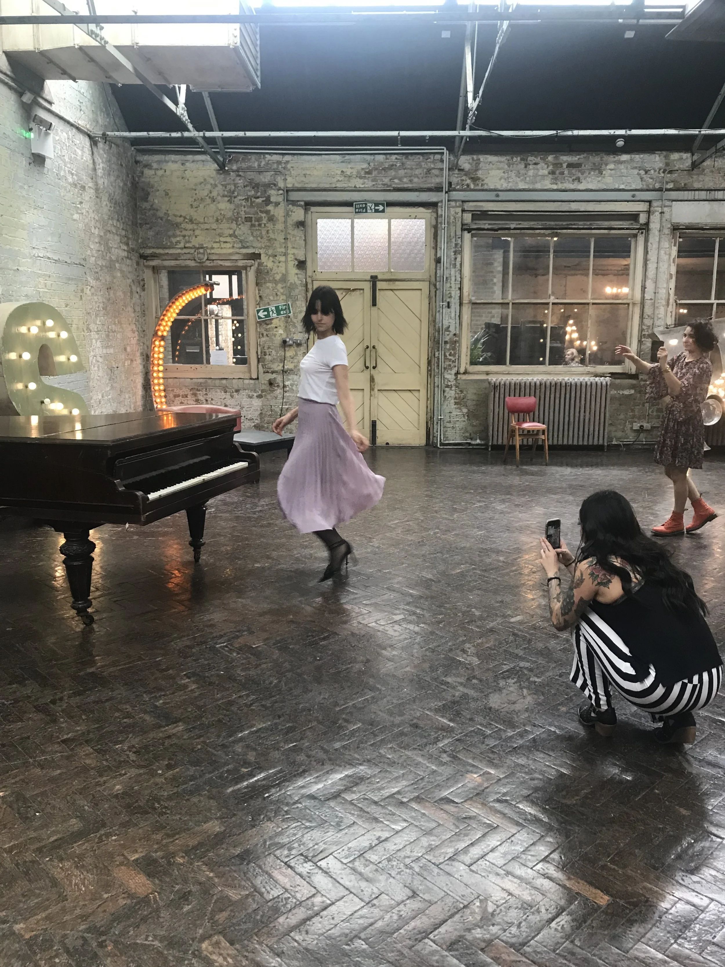A woman in a white top and purple skirt dancing near a grand piano in an industrial-style room. Another woman is crouched, taking a photo with a smartphone, wearing striped pants. A third person is in the background, gesturing. The room has exposed brick walls, large windows, and decorative light fixtures.