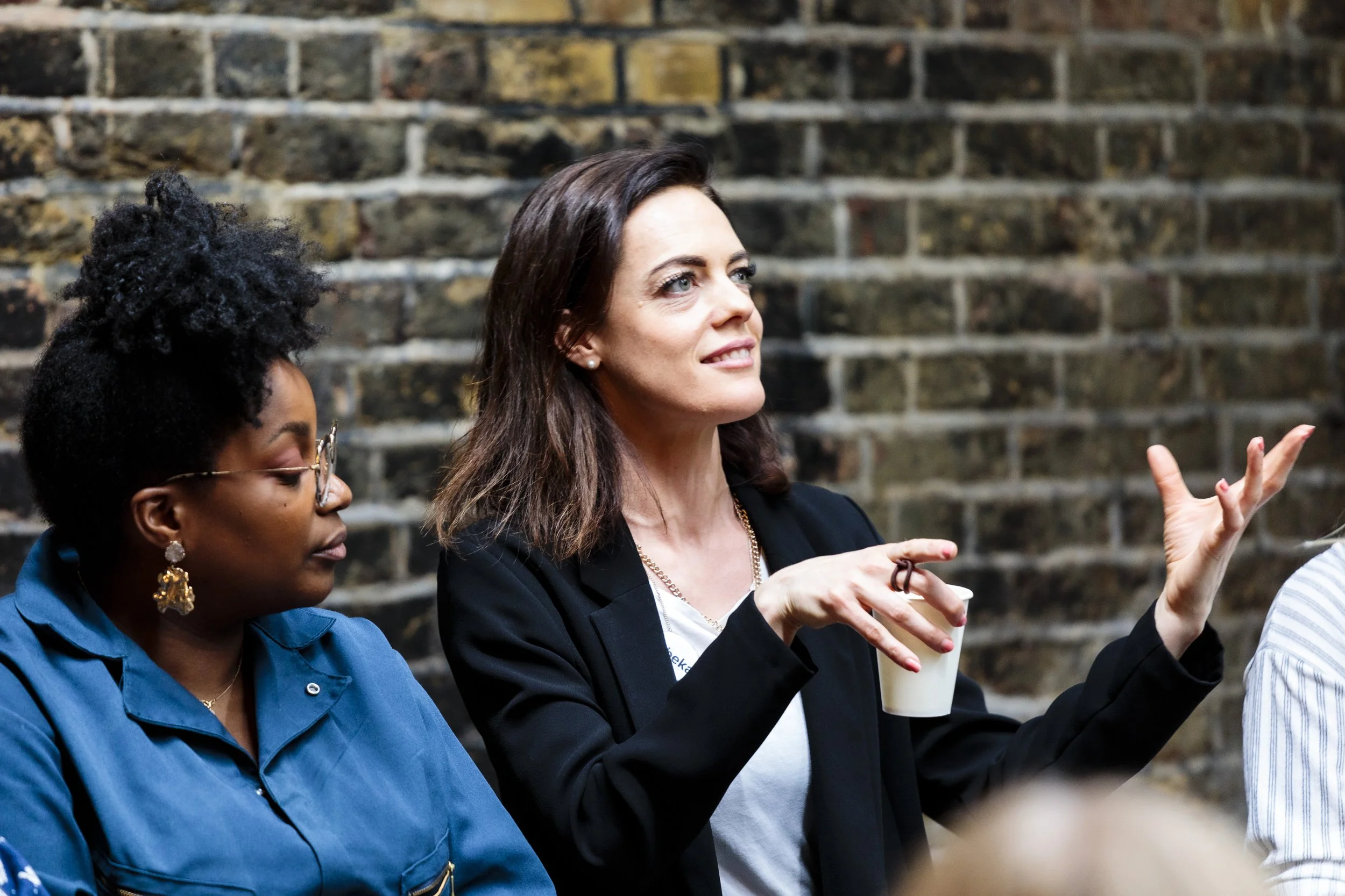 Two women sitting and talking against a brick wall; one woman is gesturing while holding a coffee cup.