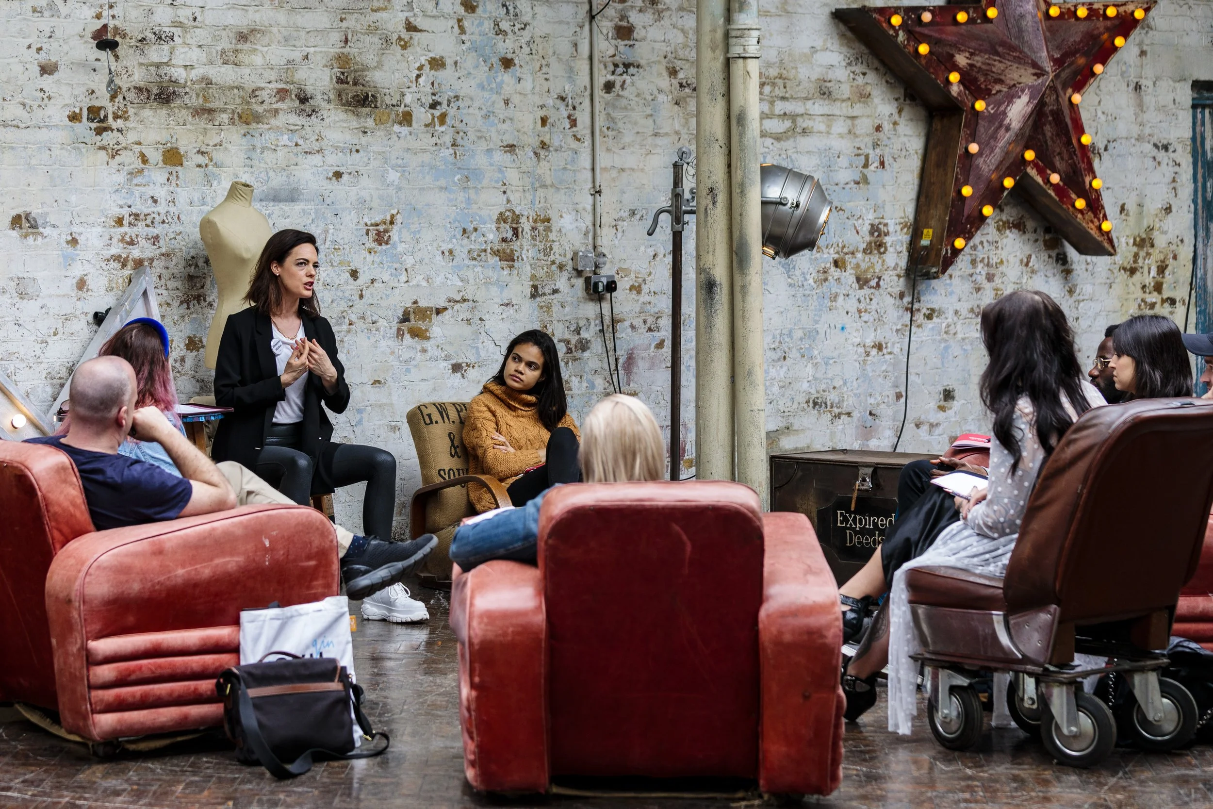 Informal group meeting in a rustic setting with mismatched armchairs, a mannequin, and a star-shaped light decoration on a brick wall.