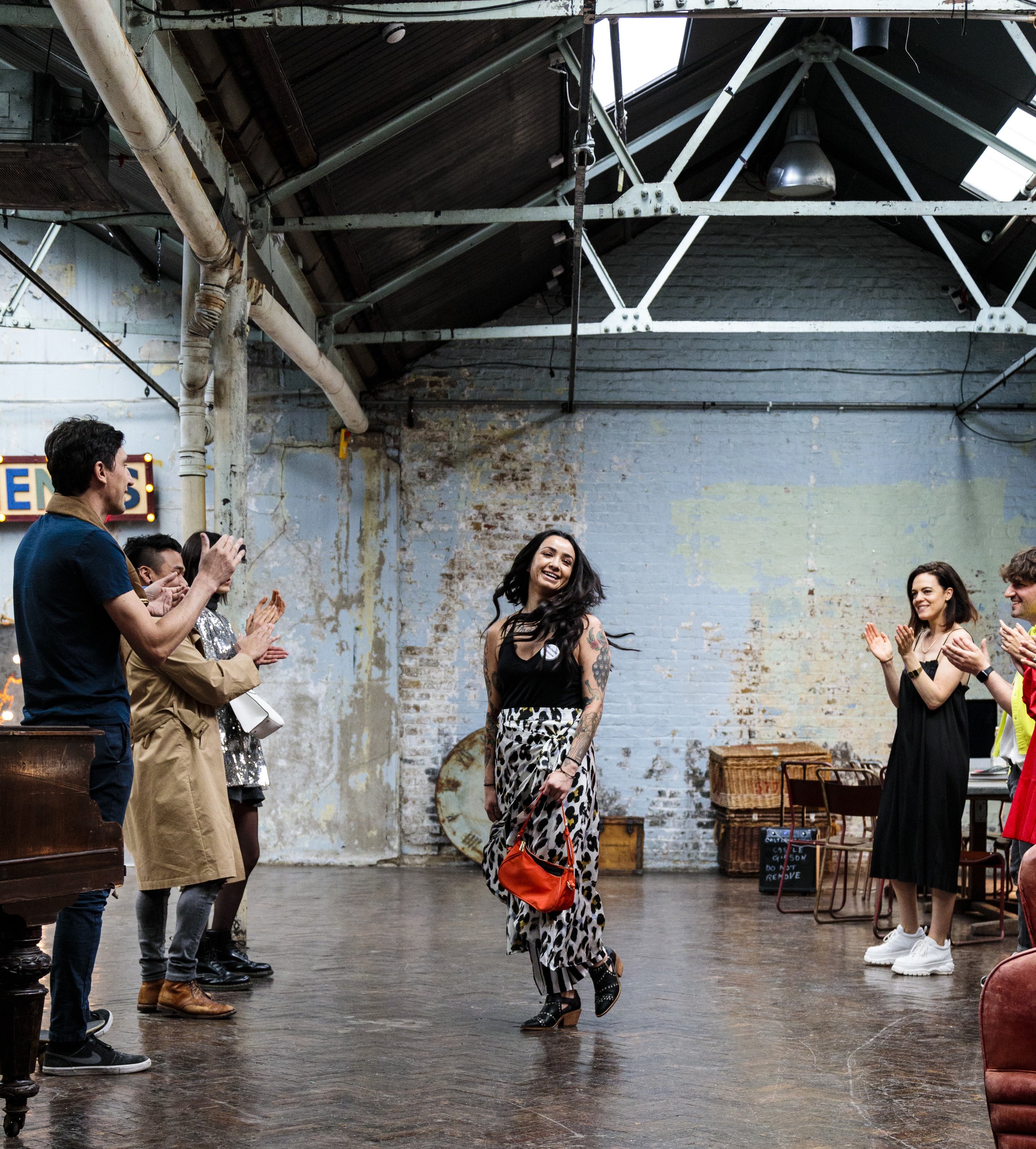 A woman in a patterned outfit walking joyfully in a rustic industrial space with an audience clapping on both sides. Exposed metal beams and a weathered brick wall create a vintage atmosphere.