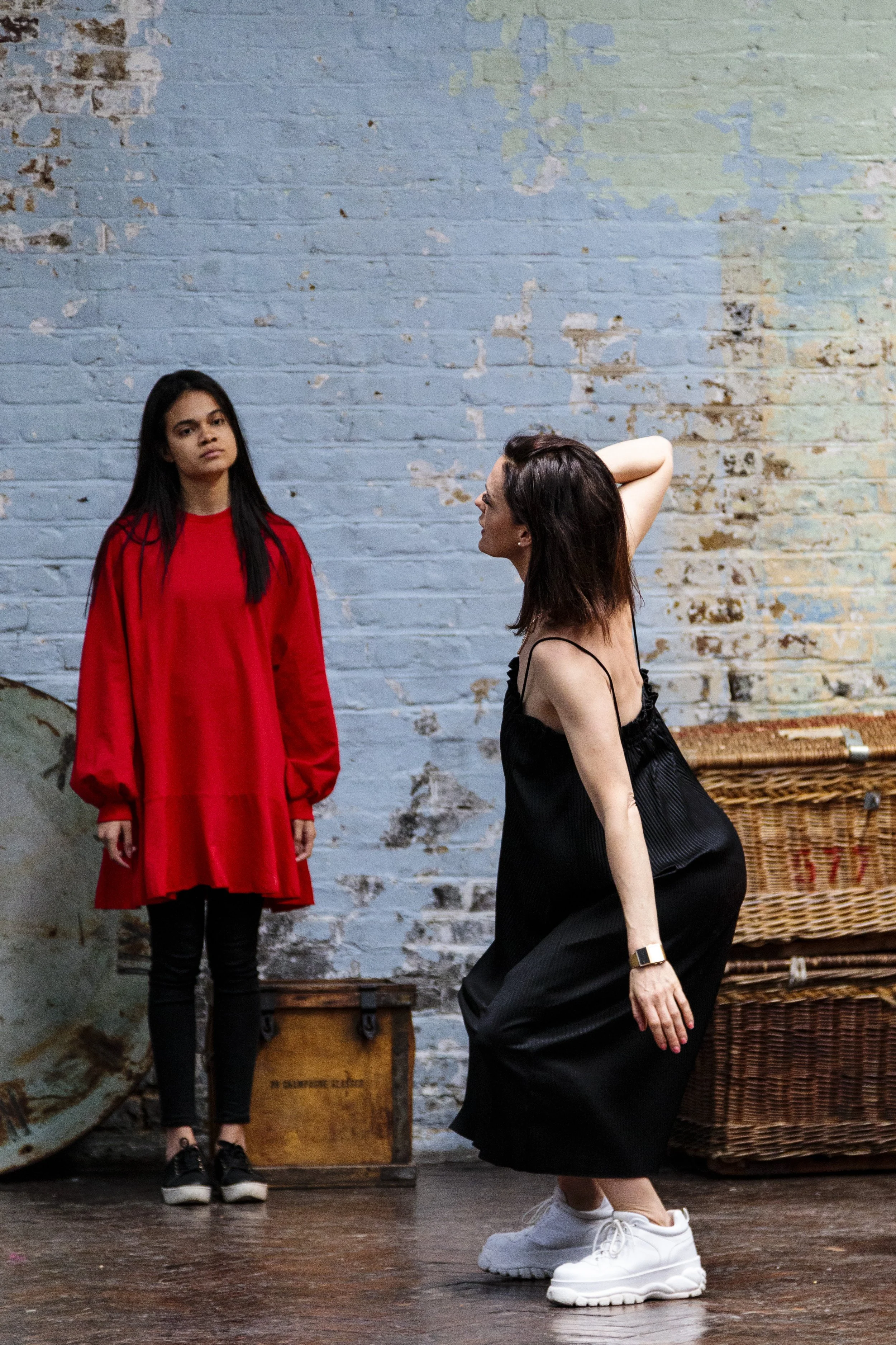 Two women in a brick-walled room, one in a red dress, the other in a black dress and sneakers, engaging in a dance or performance.