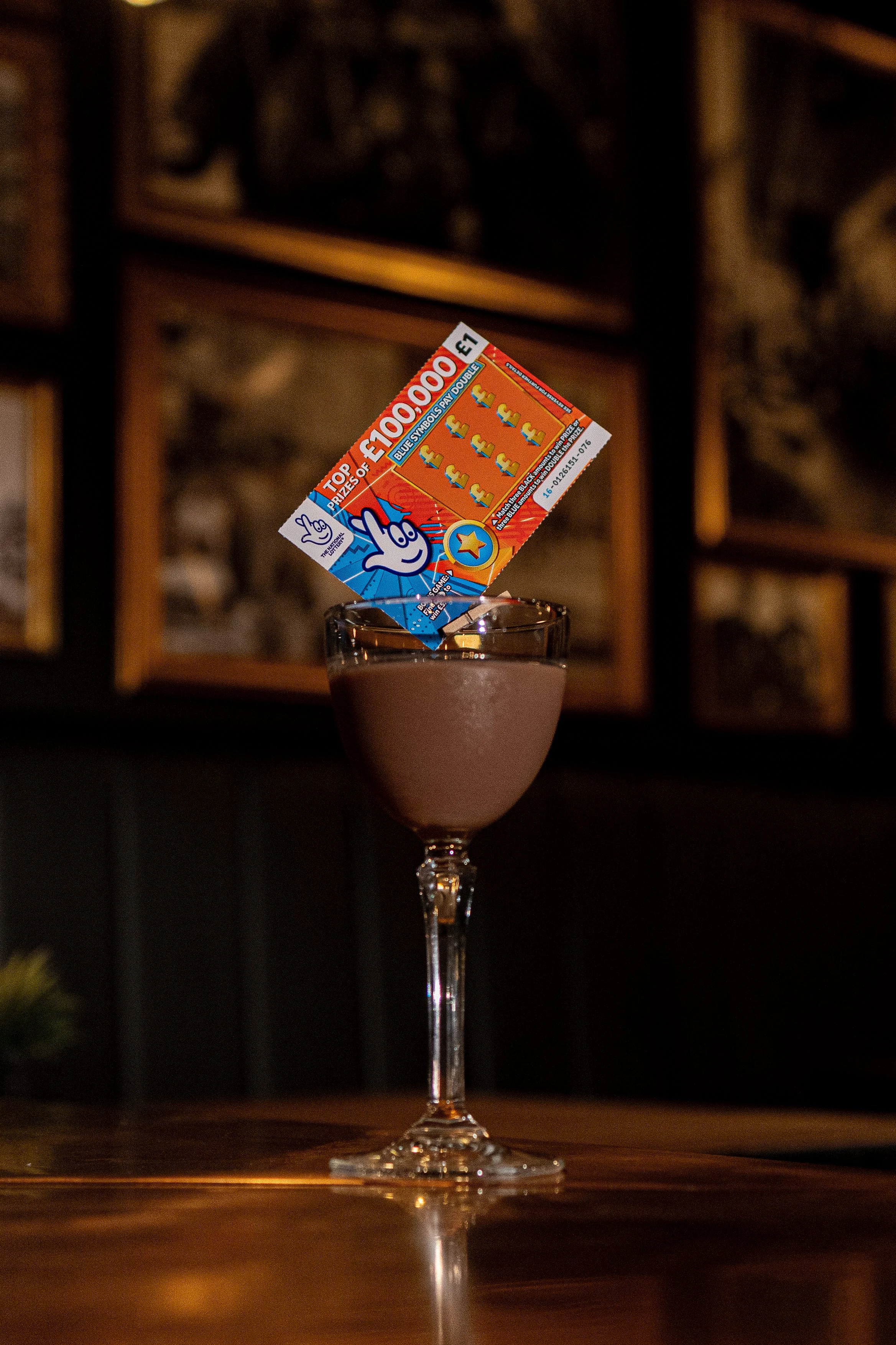 A glass of pink cocktail with a lottery ticket inserted in the glass, against a dark background with framed artwork.