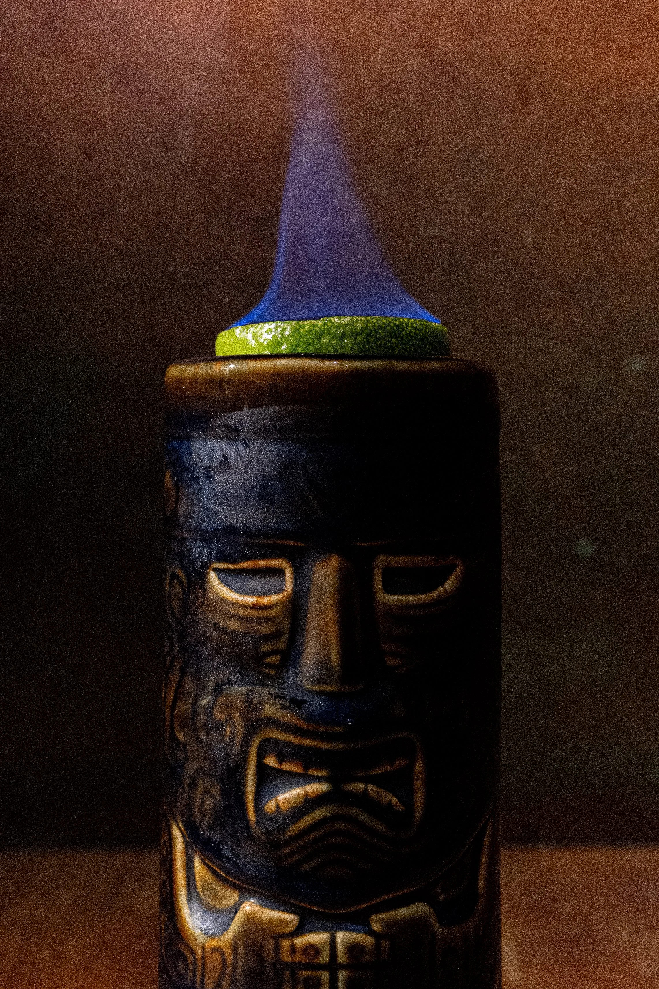 A tiki torch with a carved face design, emitting blue flame through an opening at the top, with a slice of cucumber placed around the base of the flame.