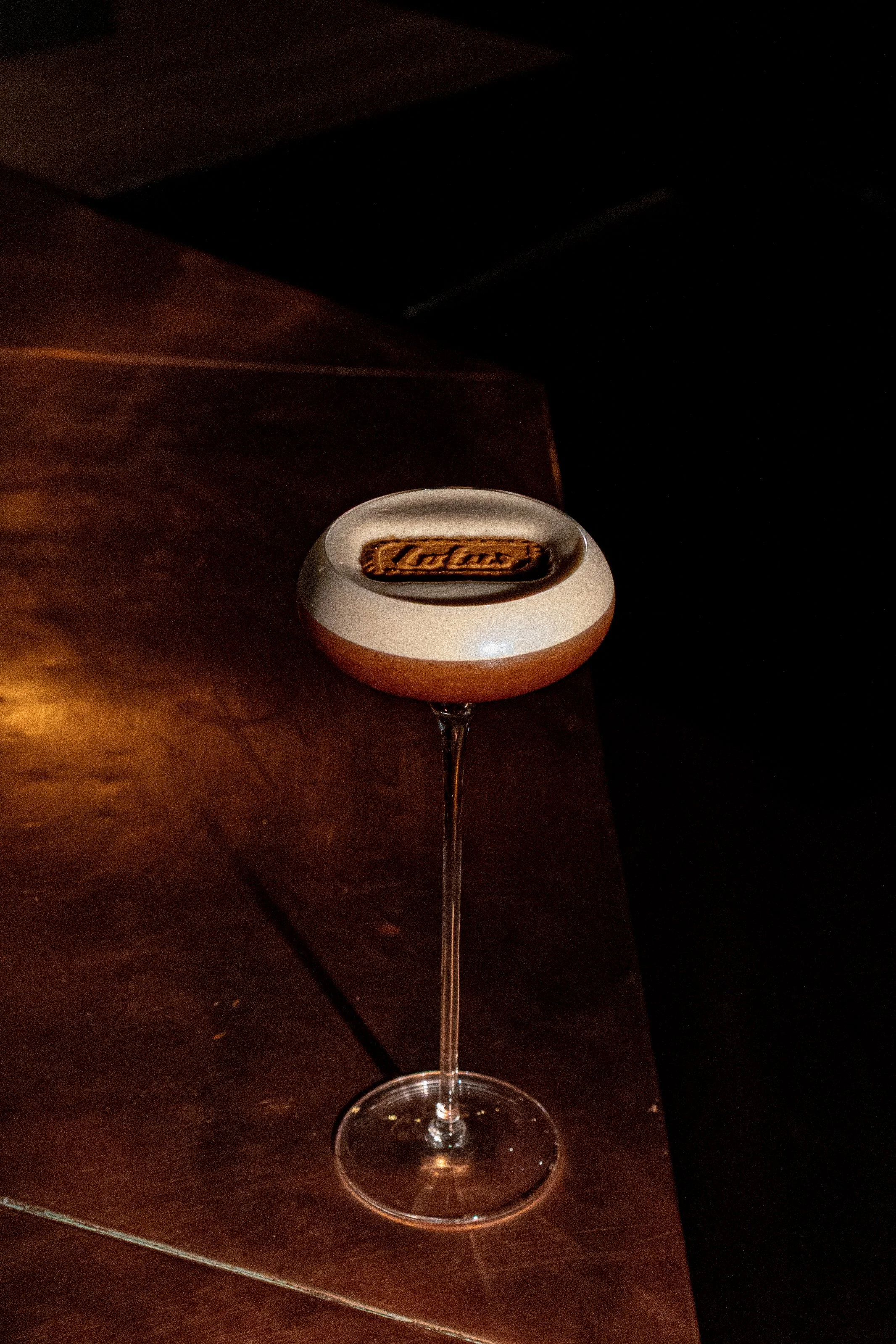 A cocktail glass with a long stem and a wide bowl, containing a layered drink topped with a cookie and a decorative garnish.